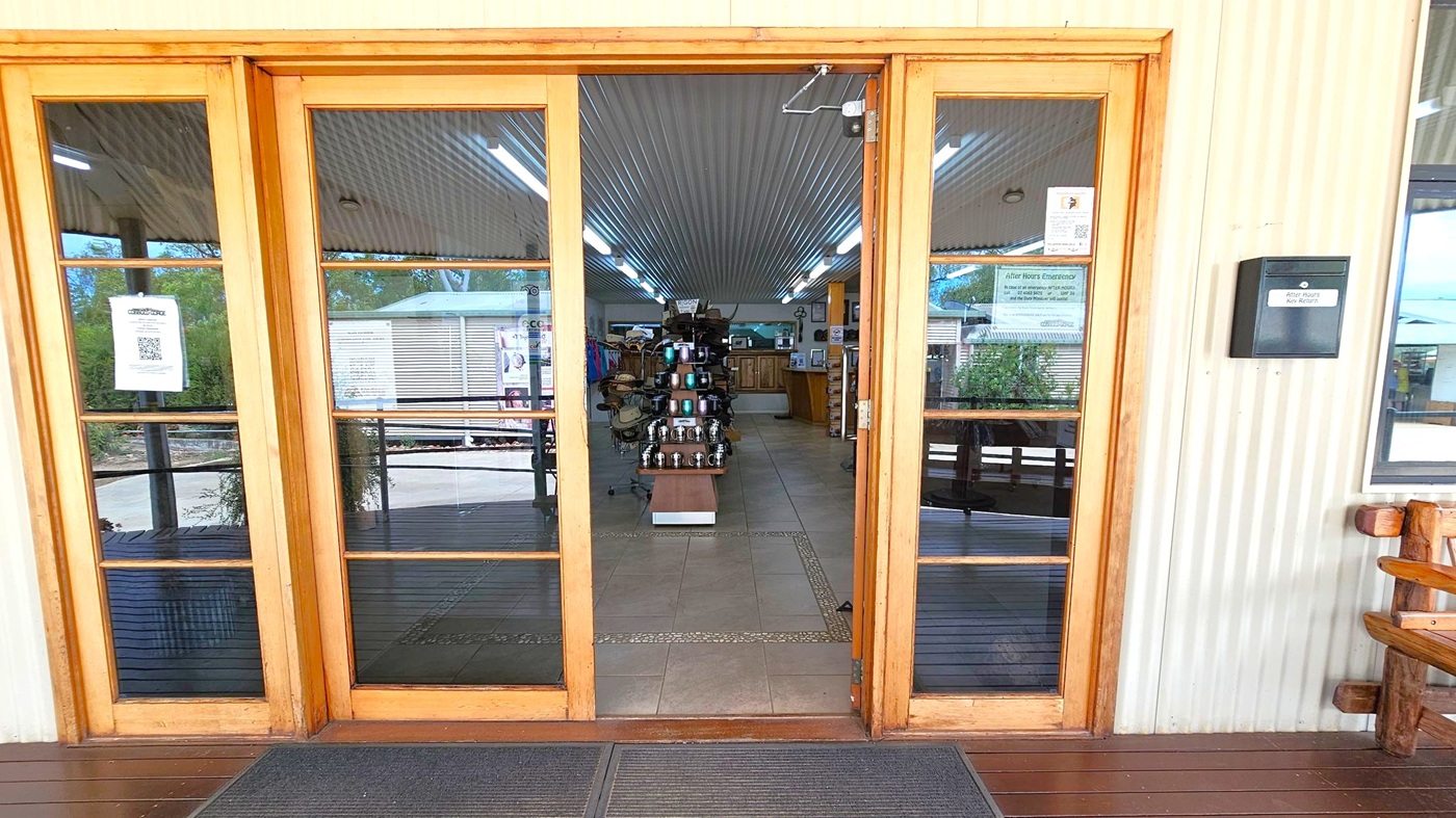 Wooden-framed glass doors lead into a shop with a corrugated metal ceiling and tiled floor. Inside, a display stand with various items is centred, surrounded by shelves and counters. Fluorescent lights line the ceiling. Outside, a wooden deck is visible, with a bench on the right. A black box labelled "After Hours Key Return" is mounted on the wall to the right of the door. Signs and notices are posted on the doors.