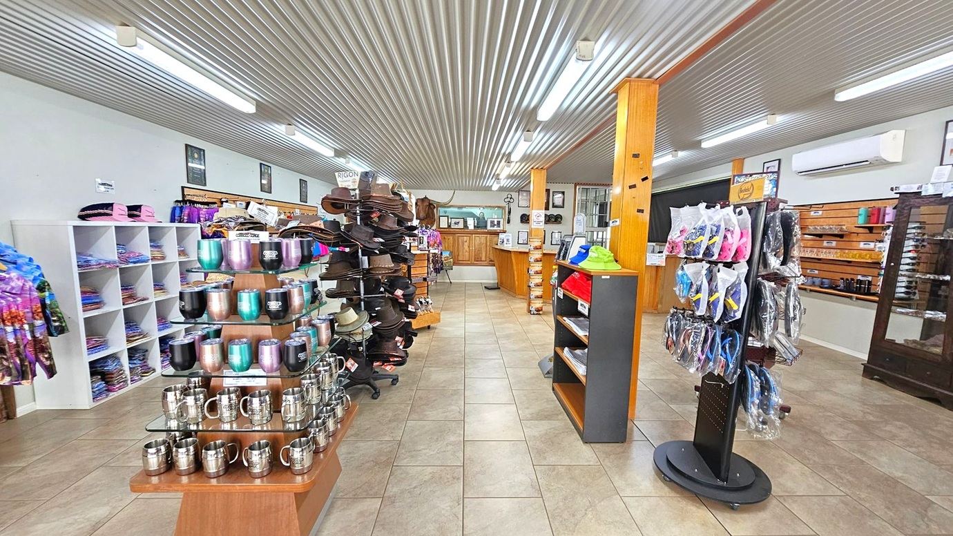 A shop interior with a corrugated metal ceiling and tiled floor. On the left, shelves display colourful clothing and a variety of mugs. In the centre, a stand holds numerous hats. To the right, a rotating rack features packaged items, and shelves display more products. The back wall has a wooden counter and decorative items. Fluorescent lights illuminate the space, and wooden pillars support the ceiling.