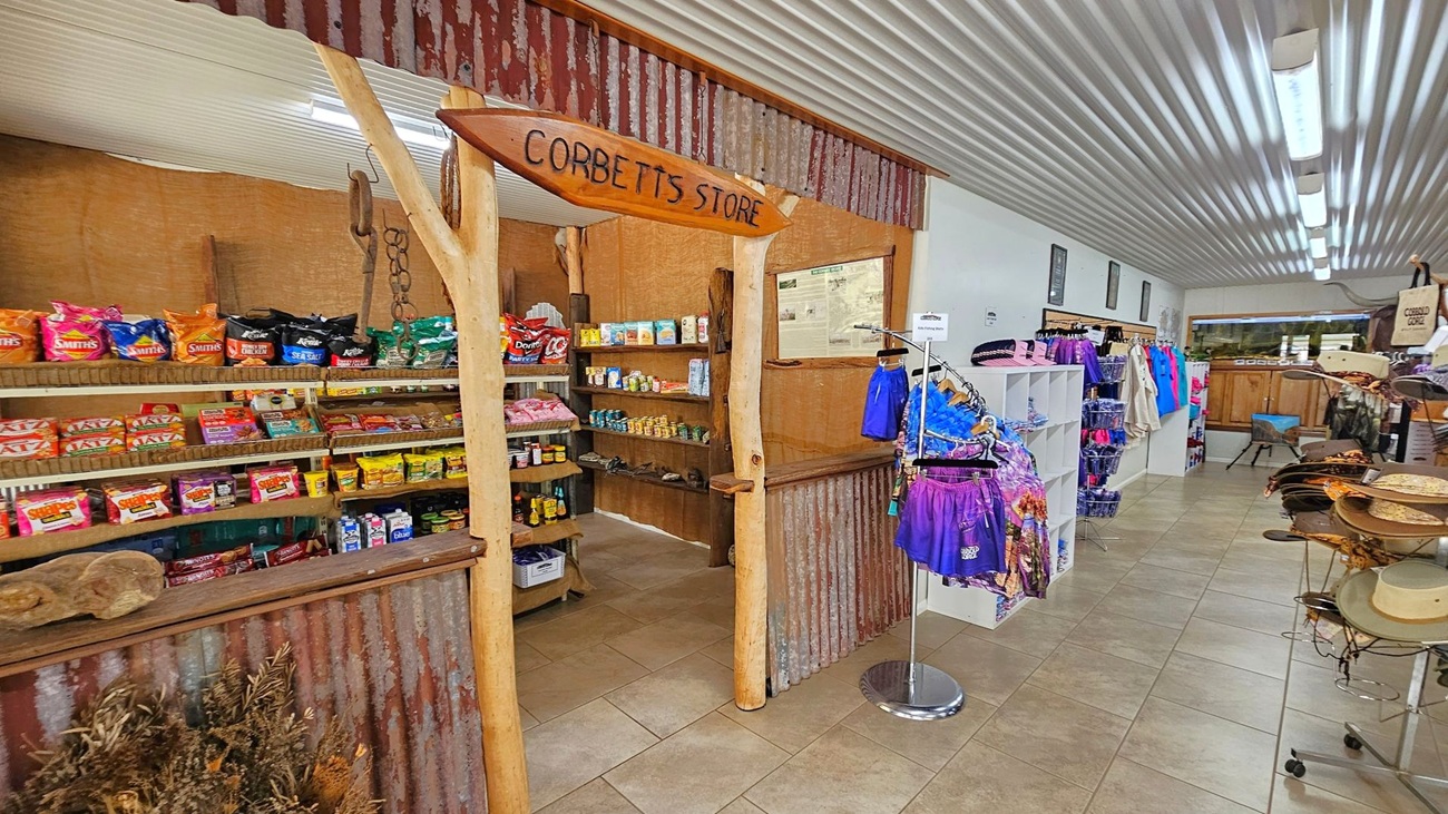 A rustic store interior with a corrugated metal and wood facade. On the left, shelves display various snacks and canned goods. In the centre, a wooden sign reads "Corbett's Store." On the right, racks hold colourful clothing and accessories. The floor is tiled, and the ceiling features corrugated metal with fluorescent lighting.