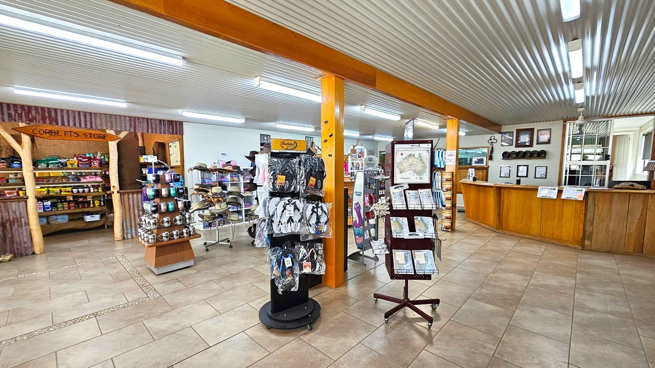A shop interior with a tiled floor and a corrugated metal ceiling. On the left, shelves display various packaged goods under a wooden sign. In the centre, rotating stands hold flip-flops and maps. To the right, a wooden counter with framed certificates and hats on the wall behind it. Fluorescent lights illuminate the space, and there are wooden beams supporting the ceiling.