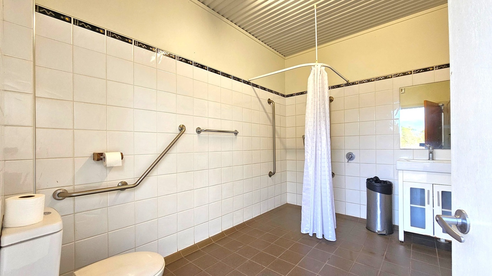 A bathroom with white tiled walls and a dark tiled floor. On the left, there's a toilet with a roll of toilet paper on top and a wall-mounted toilet paper holder. Metal grab bars are installed on the walls. In the centre, a white shower curtain hangs from a ceiling-mounted rail. On the right, a sink with a mirror above it and a stainless steel bin below. The ceiling is corrugated metal.