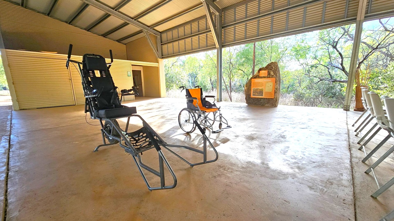 Under a metal roof, a recumbent tricycle and a wheelchair are on a concrete floor. To the right, a row of white chairs is lined up. In the background, a large rock with an informational sign stands near trees and foliage visible through the open sides of the structure. The walls are made of corrugated metal.