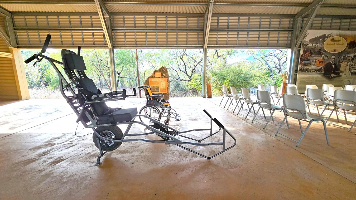 A covered outdoor area with a concrete floor. On the left, there is a specialised wheelchair with a large front wheel and handlebars, designed for off-road use. Next to it is a standard wheelchair. On the right, rows of white plastic chairs are arranged facing a wall with informational displays. The background features large windows revealing a view of trees and greenery. The ceiling is made of corrugated metal.
