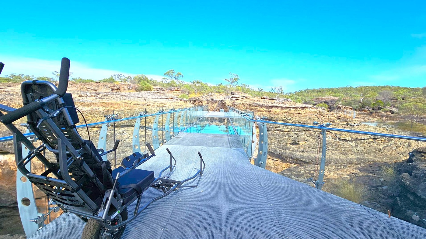 A glass-bottomed bridge extends across a rocky landscape with sparse vegetation. On the left, a black recumbent bike is parked on the bridge. The bridge has transparent railings supported by metal posts. In the background, there are rocky outcrops and scattered trees under a clear blue sky. The floor of the bridge is made of metal grating, and the glass panels reflect the sky.