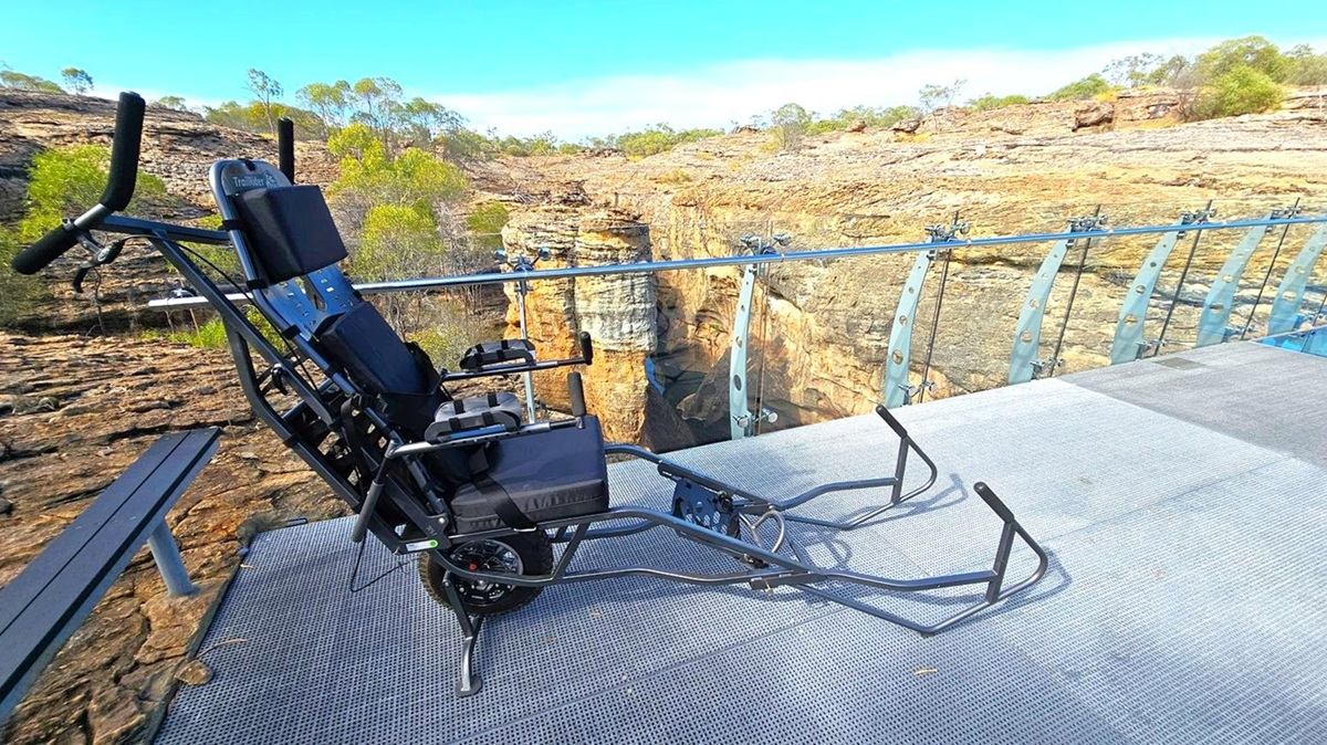 A rugged wheelchair is positioned on a metal mesh platform with a glass railing. The background features a rocky landscape with sparse vegetation and a deep gorge. The platform extends over the edge, providing a clear view of the canyon below. The wheelchair has a sturdy frame and is designed for off-road use. The surrounding area is arid, with scattered trees and shrubs on the rocky terrain.