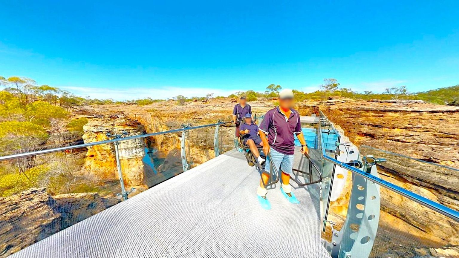 A group of three people are on a glass-bottomed walkway with metal railings, suspended over a rocky canyon. The person in front is pushing a wheelchair. The background features rugged rock formations and sparse vegetation, typical of a dry, arid landscape. The sky is clear and blue, and the walkway appears sturdy with a metal mesh floor.