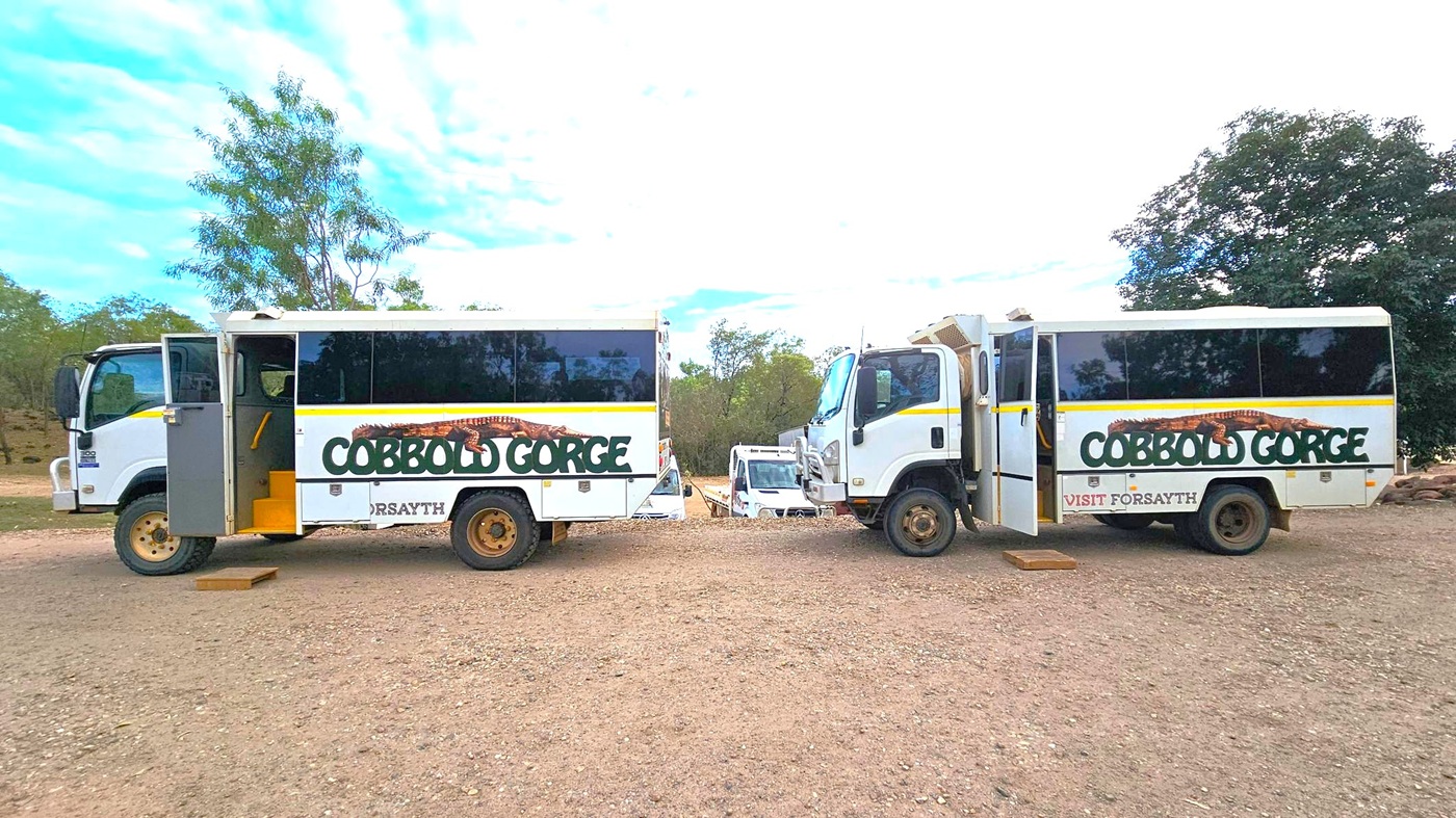 Two white tour buses with "Cobbold Gorge" written on the sides are parked on a dirt surface. The bus on the left has its door open, revealing yellow steps. Both buses have large windows and are facing each other. In the background, there are trees and several other vehicles partially visible. The sky is partly cloudy.