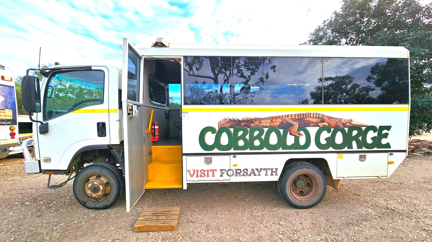 A white bus with a yellow stripe and the words "Cobbold Gorge" and "Visit Forsayth" on the side. The bus features an image of a crocodile above the text. The door is open, revealing yellow steps and a fire extinguisher inside. The bus is parked on a gravel surface, with another vehicle partially visible on the left. Trees are reflected in the bus windows and visible in the background.