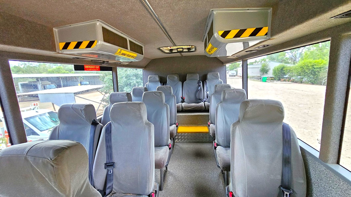 Inside a bus, there are two rows of grey upholstered seats with seatbelts, leading to a raised platform at the back with additional seating. Overhead, there are storage compartments with yellow and black caution stripes. Large windows on both sides provide a view of the outside, showing a dirt road, greenery, and some parked vehicles. The ceiling is lined with panels, and the floor is covered with a dark, textured material.