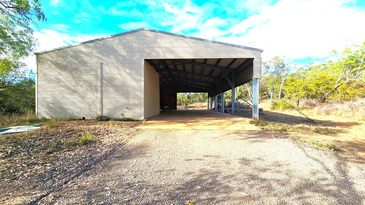 A large, open-sided metal shed with a corrugated exterior stands in the centre. The ground in front is a mix of gravel and dirt, leading into the shed. To the left, there are trees and bushes casting shadows on the shed's wall. On the right, more trees and dry grass are visible, extending into the background. The shed's roof is supported by metal beams, and the interior is partially visible, showing more trees through the back opening.