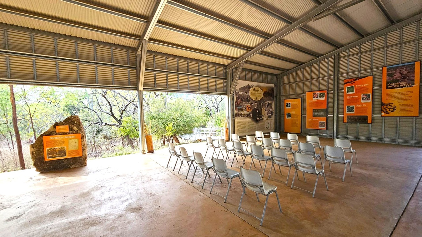 An open-air pavilion with a corrugated metal roof and walls. On the right, several informational panels with text and images are mounted on the wall. In the centre, rows of white plastic chairs are arranged facing the panels. On the left, a large rock with an informational sign stands near the open side of the pavilion, revealing a view of trees and foliage outside. The floor is concrete, and the ceiling is metal.