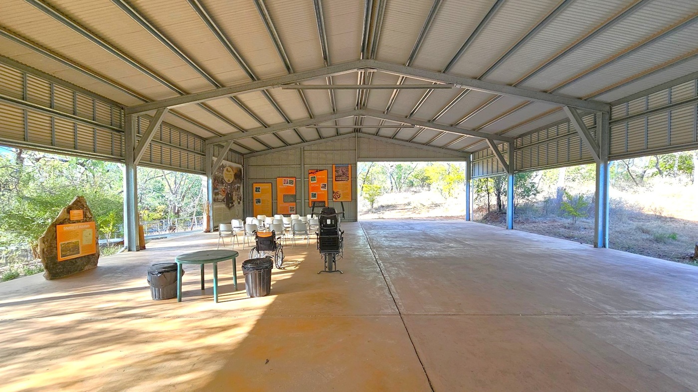 A large open-sided structure with a metal roof and beams. On the left, there is a rock with an informational sign and a few trees visible outside. In the centre, several white chairs are arranged in rows facing a wall with orange informational boards. A wheelchair and a small table with bins are also present. The right side opens to a view of more trees and grass. The floor is concrete, and the ceiling is made of corrugated metal.