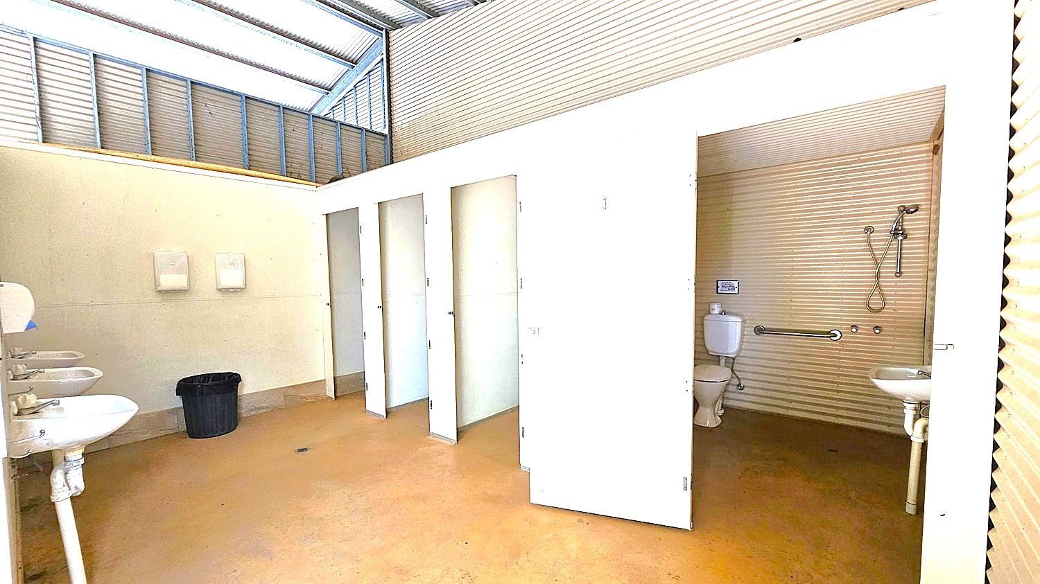 A public restroom with a corrugated metal ceiling and walls. On the left, there are three white sinks with mirrors above them and a black waste bin on the floor. In the centre, there are three closed white stall doors and one open door revealing a toilet, a grab bar, and a small sink. The floor is a light brown colour, and the upper walls have a light green hue. The ceiling has a slanted design with translucent panels allowing natural light.