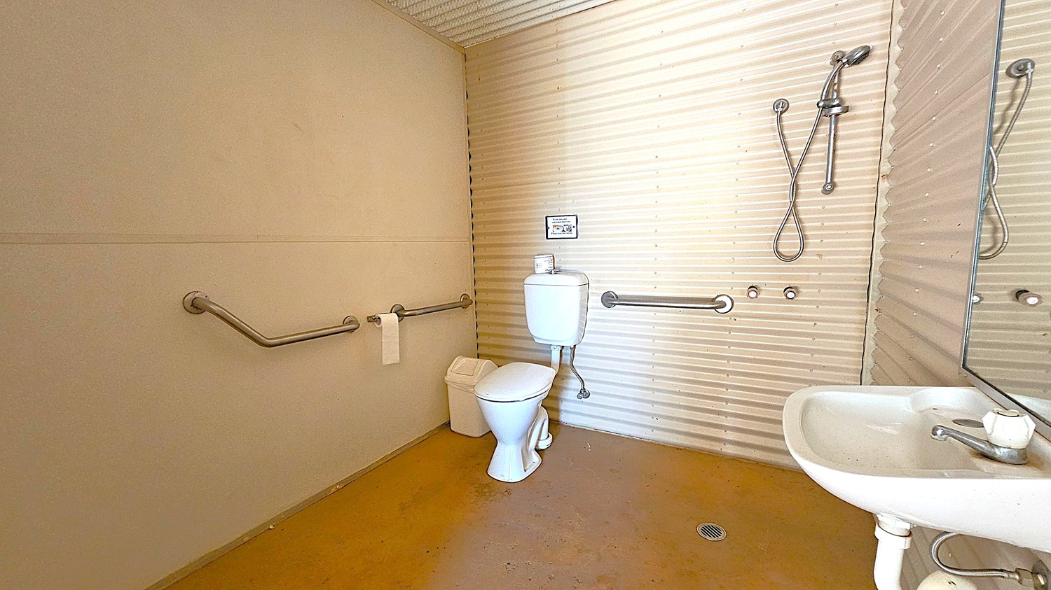 A bathroom with corrugated metal walls and a brown floor. On the left, there are metal grab bars and a toilet with a white cistern. A small bin is placed beside the toilet. On the right, a white sink with a single tap is mounted on the wall, accompanied by a mirror. Above the sink, a handheld showerhead is attached to the wall. The ceiling is also corrugated metal.
