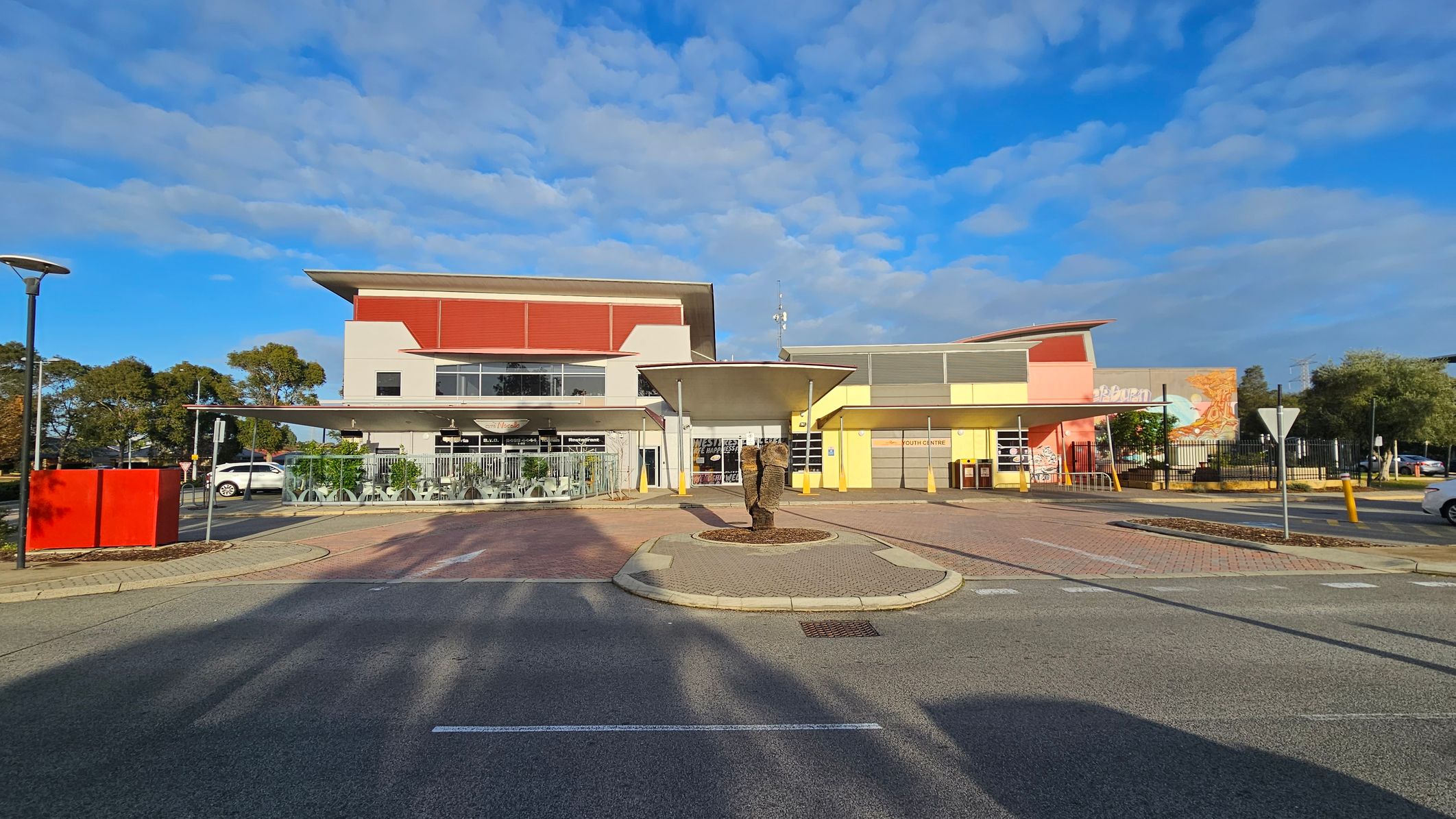 A modern building with a flat roof and a mix of red, yellow, and grey colours. In the centre foreground, there's a small roundabout with a sculpture. The left side has a glass-enclosed outdoor seating area with tables and chairs. On the right, a yellow section of the building with a sign reading "Youth Centre" and a mural. The ground is paved with a mix of asphalt and brick patterns. Trees and parked cars are visible in the background. A modern building with a flat roof and a mix of red, yellow, and grey colours. In the centre foreground, there's a small roundabout with a sculpture. The left side has a glass-enclosed outdoor seating area with tables and chairs. On the right, a yellow section of the building with a sign reading "Youth Centre" and a mural. The ground is paved with a mix of asphalt and brick patterns. Trees and parked cars are visible in the background.