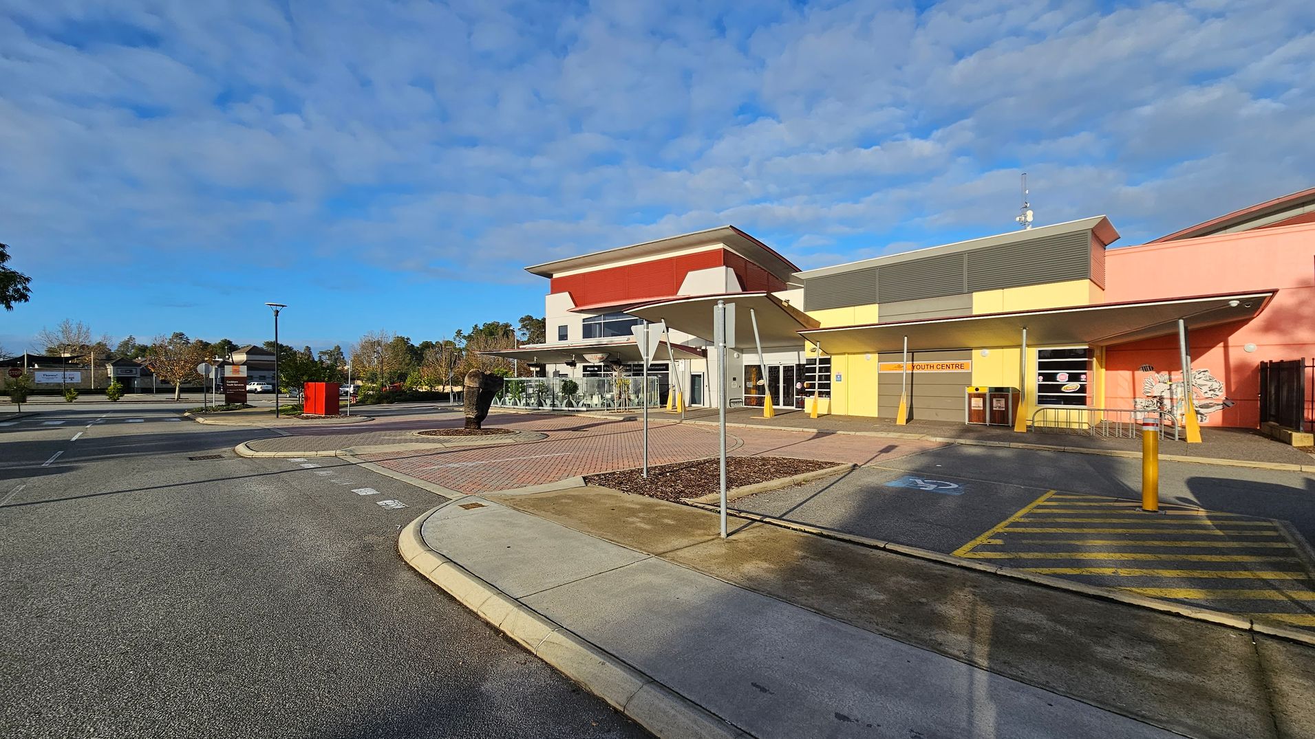 A modern building with a red and white facade is on the left, featuring large windows and a shaded outdoor area. To the right, a yellow and pink structure labelled "Youth Centre" has a sloped roof and several windows. In the foreground, a paved area with a roundabout and a sculpture is visible, along with a pedestrian crossing marked in yellow. The background includes trees and a few parked cars. The ground is a mix of concrete and asphalt. A modern building with a red and white facade is on the left, featuring large windows and a shaded outdoor area. To the right, a yellow and pink structure labelled "Youth Centre" has a sloped roof and several windows. In the foreground, a paved area with a roundabout and a sculpture is visible, along with a pedestrian crossing marked in yellow. The background includes trees and a few parked cars. The ground is a mix of concrete and asphalt.