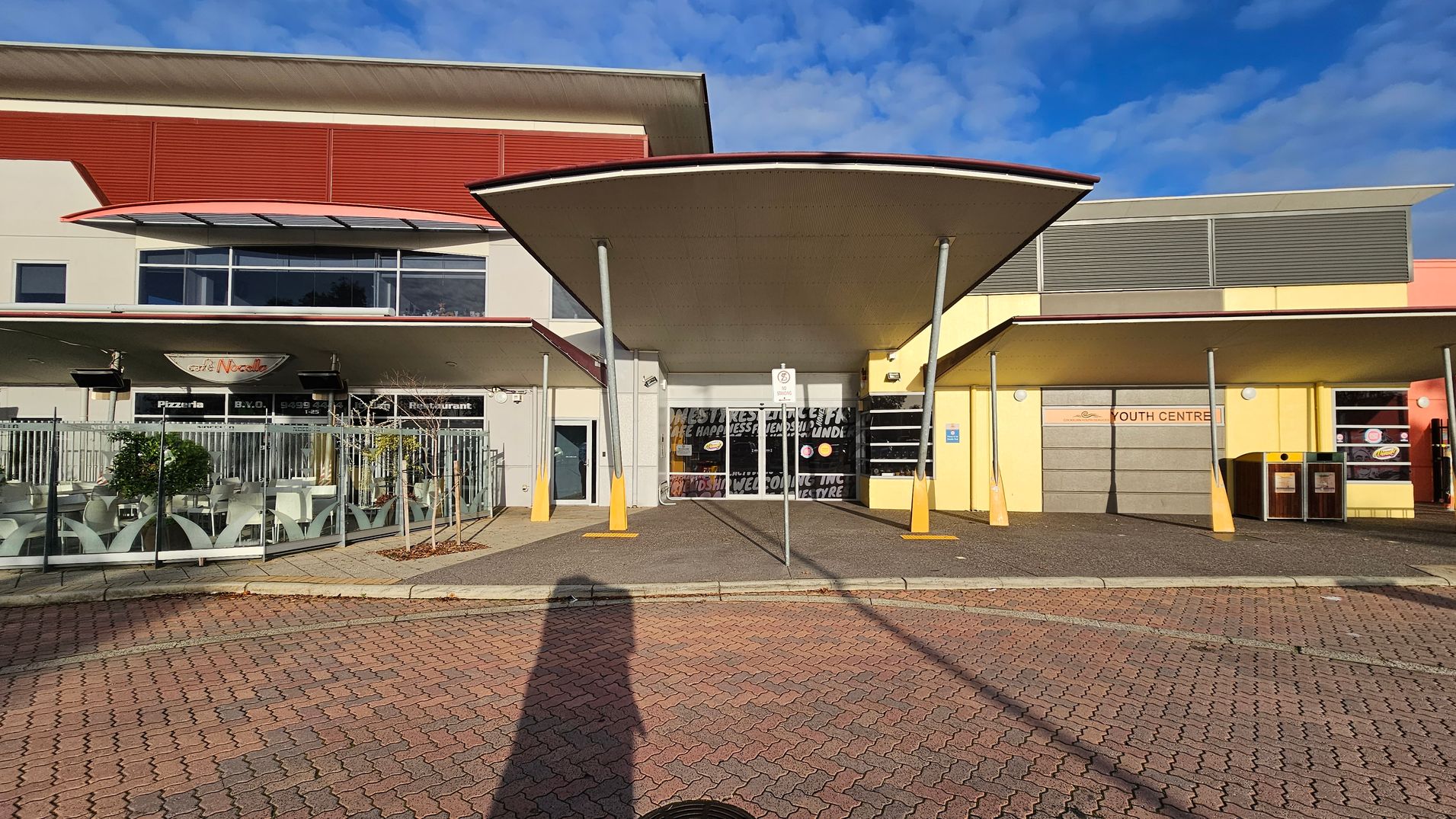 In the foreground, a paved area with a shadow cast on it. The middle features a building with a central entrance under a large, curved canopy supported by poles. To the left, a café with outdoor seating enclosed by a metal fence. To the right, a section marked "Youth Centre" with a yellow facade and two bins. The building has a mix of red, yellow, and grey panels, with windows and signage. In the foreground, a paved area with a shadow cast on it. The middle features a building with a central entrance under a large, curved canopy supported by poles. To the left, a café with outdoor seating enclosed by a metal fence. To the right, a section marked "Youth Centre" with a yellow facade and two bins. The building has a mix of red, yellow, and grey panels, with windows and signage.