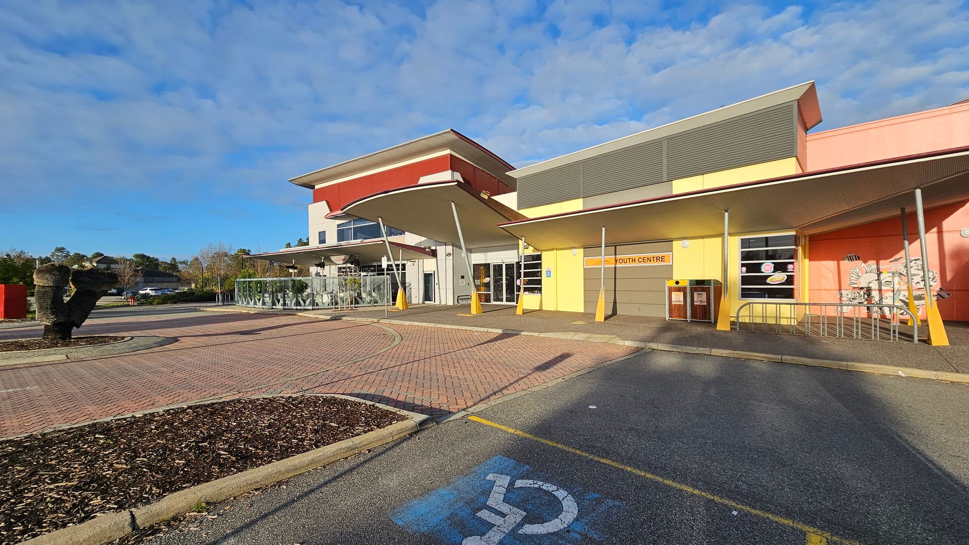 A modern building with a colourful facade, featuring yellow and orange walls, and a sign reading "Youth Centre" above the entrance. The structure has a distinctive overhanging roof supported by angled poles. In the foreground, there is a paved area with a designated disabled parking space marked in blue. To the left, a landscaped area with mulch and a sculptural element. The background includes trees and a clear sky. A modern building with a colourful facade, featuring yellow and orange walls, and a sign reading "Youth Centre" above the entrance. The structure has a distinctive overhanging roof supported by angled poles. In the foreground, there is a paved area with a designated disabled parking space marked in blue. To the left, a landscaped area with mulch and a sculptural element. The background includes trees and a clear sky.