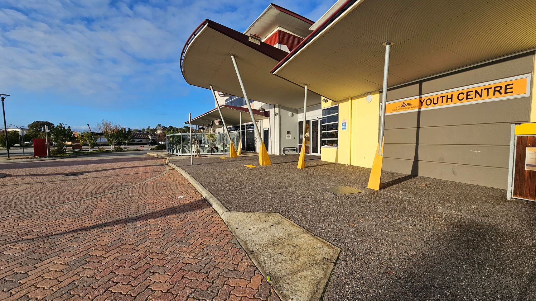 A building with a sign reading "Youth Centre" on the right, featuring a yellow and grey facade. The structure has a modern design with slanted pillars supporting overhanging roofs. The foreground shows a paved area with a mix of concrete and brick patterns. On the left, there are trees and a few street lamps along a pathway. The background includes more trees and distant buildings under a partly cloudy sky. A building with a sign reading "Youth Centre" on the right, featuring a yellow and grey facade. The structure has a modern design with slanted pillars supporting overhanging roofs. The foreground shows a paved area with a mix of concrete and brick patterns. On the left, there are trees and a few street lamps along a pathway. The background includes more trees and distant buildings under a partly cloudy sky.