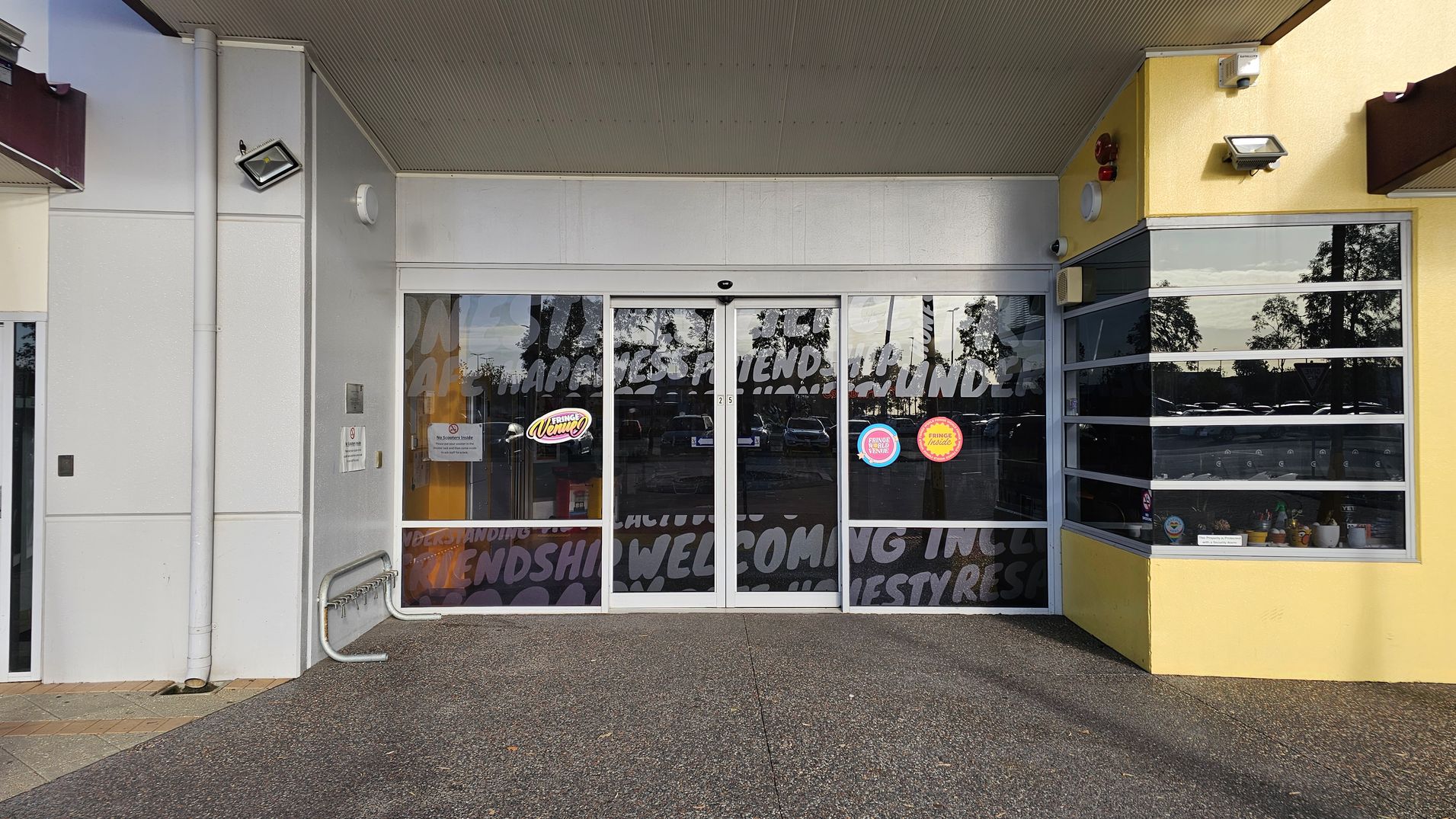 A storefront with large glass sliding doors in the centre, featuring text and logos. To the left, a white wall with a mounted light and a bike rack. On the right, a yellow wall with a window display showing various small items. The ceiling is green, and the floor is a speckled concrete surface. A storefront with large glass sliding doors in the centre, featuring text and logos. To the left, a white wall with a mounted light and a bike rack. On the right, a yellow wall with a window display showing various small items. The ceiling is green, and the floor is a speckled concrete surface.