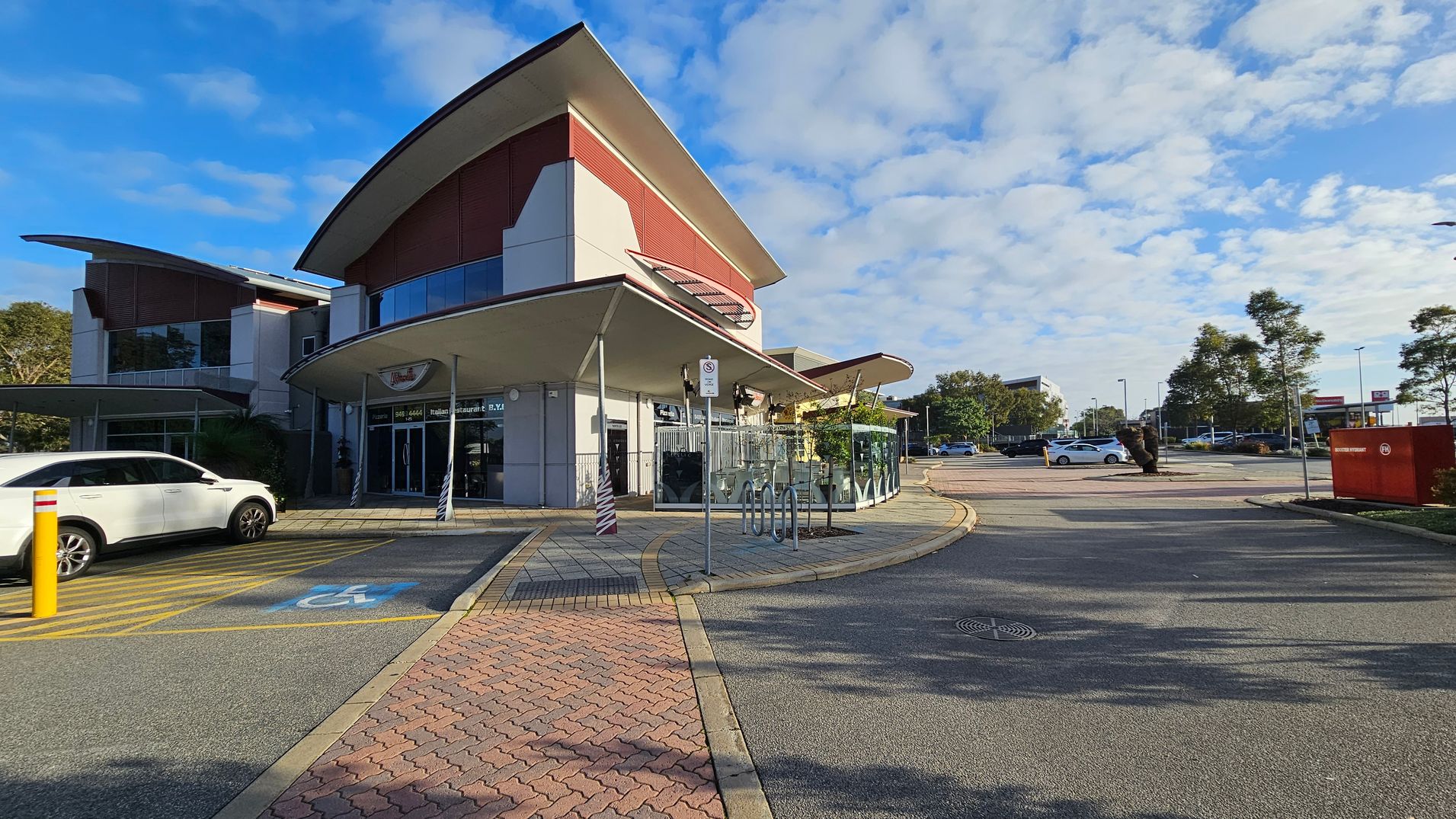 A modern building with a curved roof is on the left, featuring large glass windows and a red accent wall. A white car is parked in a designated disabled parking space with blue and yellow markings. In the centre, there is a bicycle rack and a signpost. The right side shows a spacious car park with a few trees and vehicles. The ground is paved with a mix of asphalt and brick patterns. A modern building with a curved roof is on the left, featuring large glass windows and a red accent wall. A white car is parked in a designated disabled parking space with blue and yellow markings. In the centre, there is a bicycle rack and a signpost. The right side shows a spacious car park with a few trees and vehicles. The ground is paved with a mix of asphalt and brick patterns.