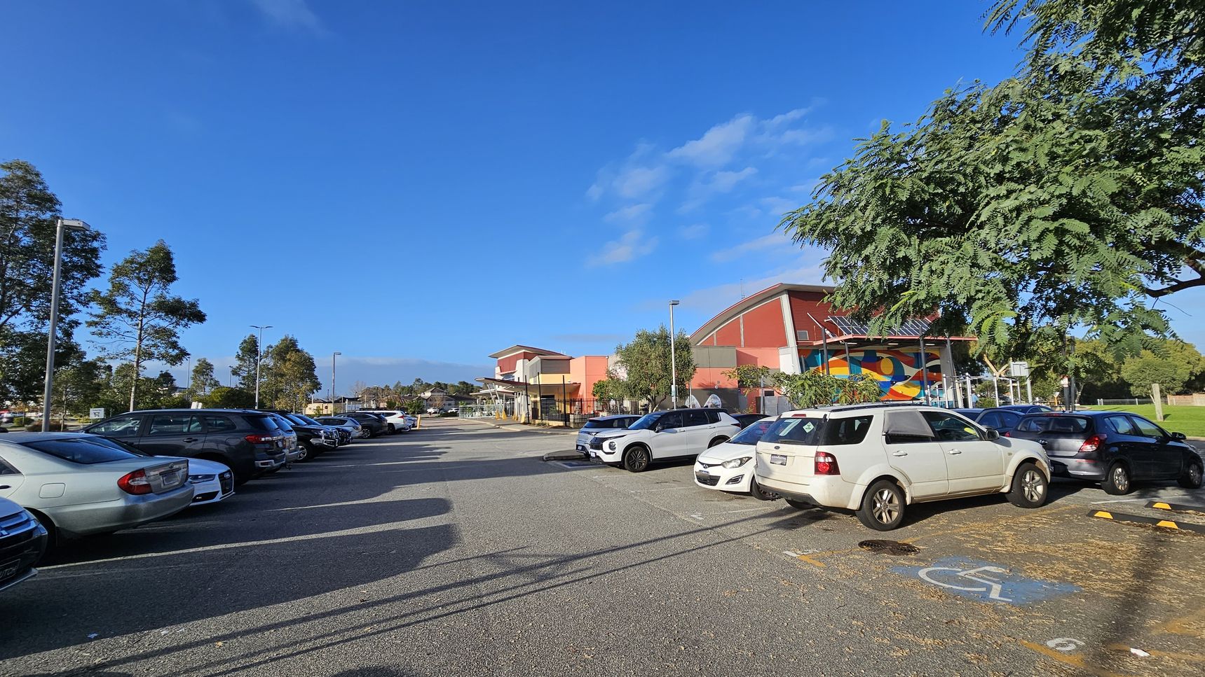A car park with several vehicles is on the left, including a white SUV in the foreground. On the right, a building with a colourful mural and a red roof is partially visible, surrounded by trees. The ground is paved, and there is a marked disabled parking space in the foreground. Tall streetlights and more trees are in the background under a clear blue sky. A car park with several vehicles is on the left, including a white SUV in the foreground. On the right, a building with a colourful mural and a red roof is partially visible, surrounded by trees. The ground is paved, and there is a marked disabled parking space in the foreground. Tall streetlights and more trees are in the background under a clear blue sky.
