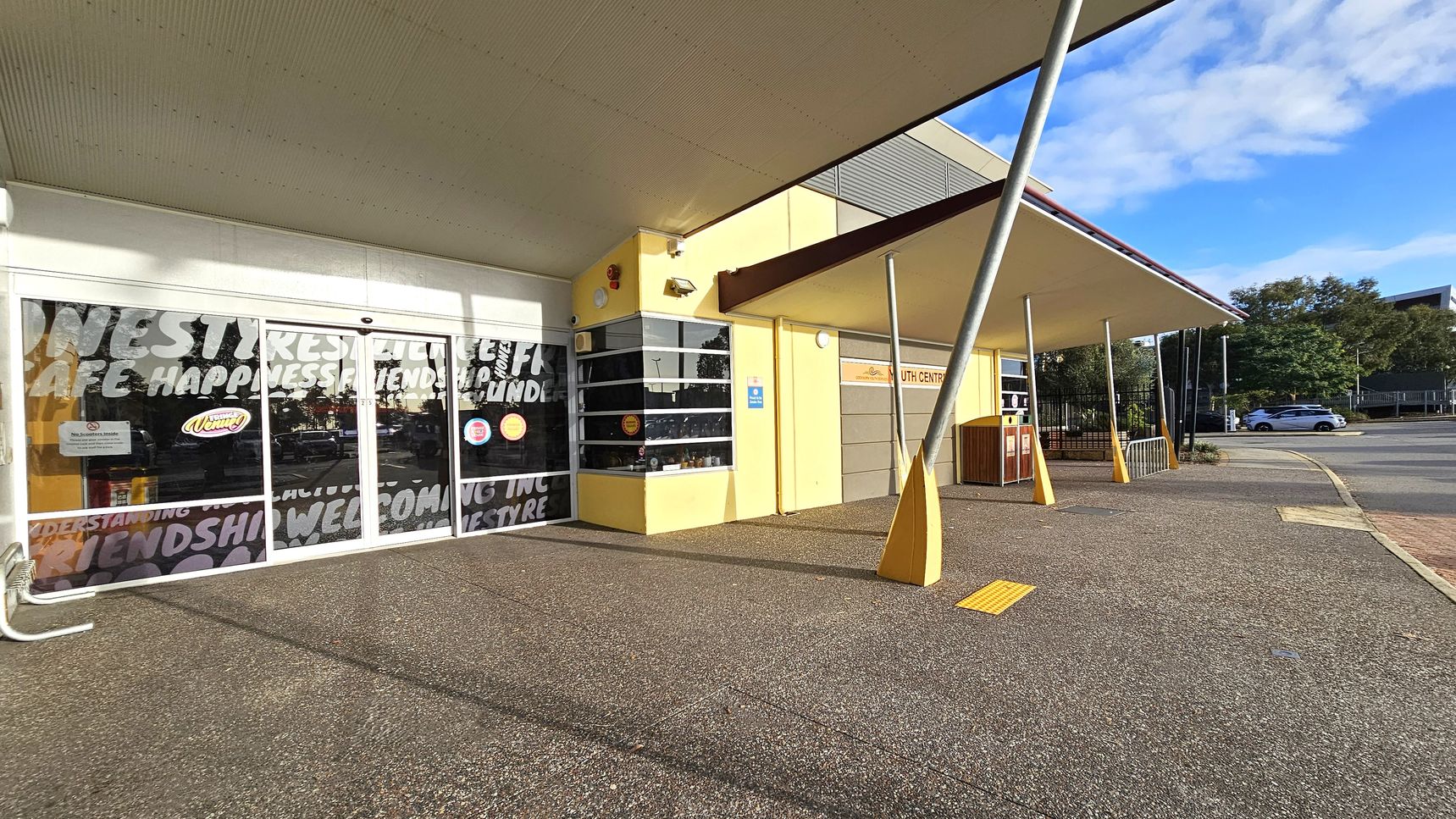 A building with a yellow exterior and a large overhanging roof. On the left, there are glass doors with words like "happiness" and "friendship" printed on them. The right side features a sign for a youth centre and a series of angled metal poles supporting the roof. The ground is paved with a textured surface, and a yellow tactile paving strip is visible. In the background, there are trees and a parked car on the street. A building with a yellow exterior and a large overhanging roof. On the left, there are glass doors with words like "happiness" and "friendship" printed on them. The right side features a sign for a youth centre and a series of angled metal poles supporting the roof. The ground is paved with a textured surface, and a yellow tactile paving strip is visible. In the background, there are trees and a parked car on the street.