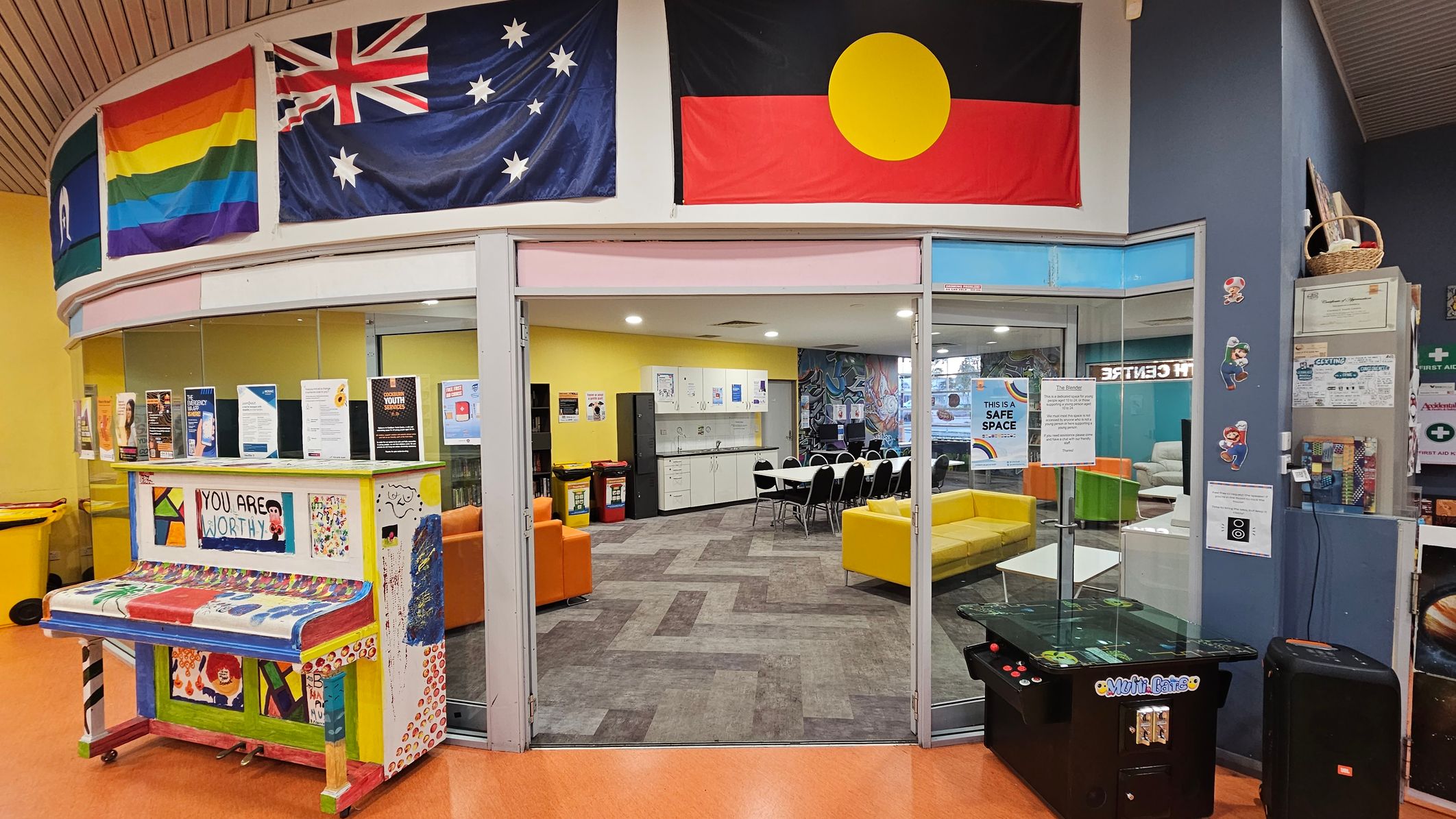 A colourful indoor space with a curved ceiling. On the left, a vibrant piano with "YOU ARE WORTHY" painted on it is surrounded by various posters. Above, flags including a rainbow flag and the Australian flag are displayed. In the centre, a glass entrance leads to a room with yellow and orange seating, a table with chairs, and a kitchenette. On the right, a gaming machine and wall decorations featuring characters are visible. The floor is a mix of orange and patterned tiles. A colourful indoor space with a curved ceiling. On the left, a vibrant piano with "YOU ARE WORTHY" painted on it is surrounded by various posters. Above, flags including a rainbow flag and the Australian flag are displayed. In the centre, a glass entrance leads to a room with yellow and orange seating, a table with chairs, and a kitchenette. On the right, a gaming machine and wall decorations featuring characters are visible. The floor is a mix of orange and patterned tiles.