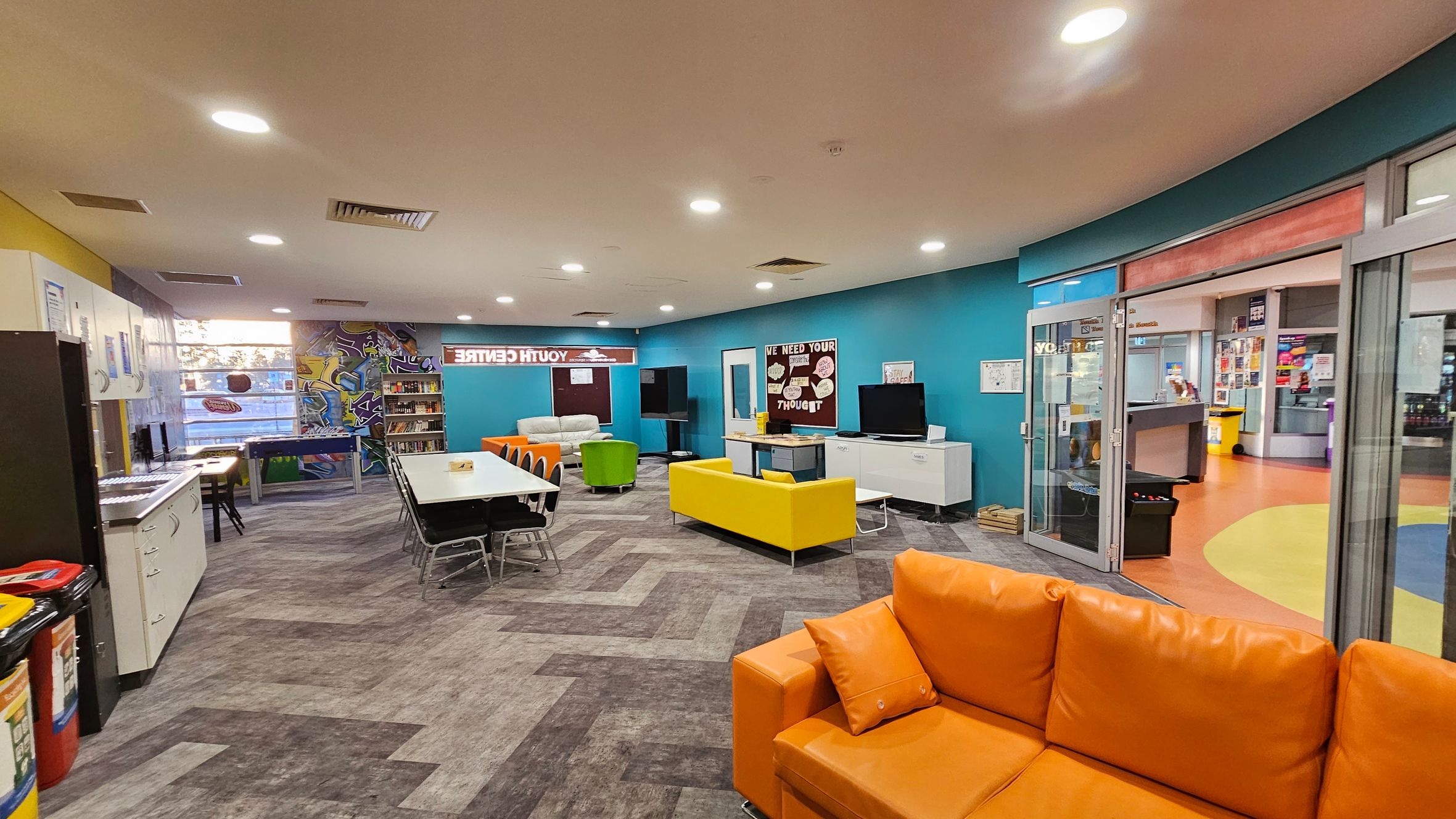 A spacious room with a vibrant, modern design. On the left, there are white cabinets and a row of computers. In the centre, a long table with black chairs is surrounded by colourful seating, including a yellow sofa and an orange couch. The back wall features a graffiti-style mural and bookshelves. On the right, glass doors lead to another area with an orange and yellow floor. The ceiling is fitted with recessed lighting, and the floor has a grey, patterned design. A spacious room with a vibrant, modern design. On the left, there are white cabinets and a row of computers. In the centre, a long table with black chairs is surrounded by colourful seating, including a yellow sofa and an orange couch. The back wall features a graffiti-style mural and bookshelves. On the right, glass doors lead to another area with an orange and yellow floor. The ceiling is fitted with recessed lighting, and the floor has a grey, patterned design.