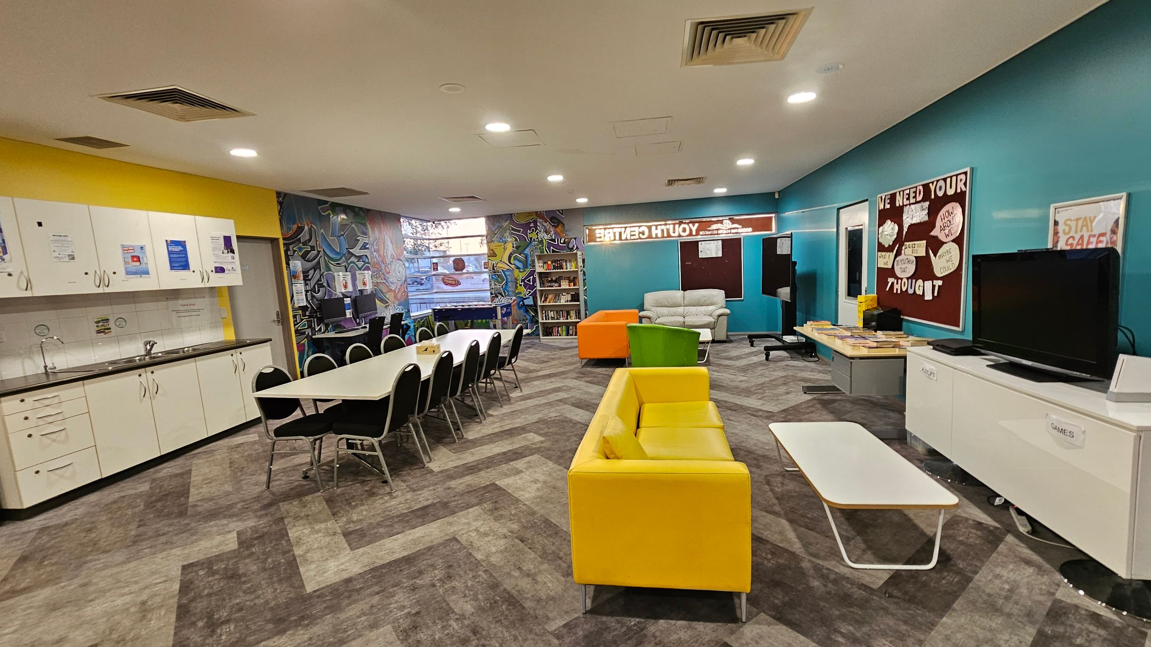 A brightly lit room with a yellow and blue colour scheme. On the left, there is a kitchenette with white cabinets and a sink. In the centre, a long table with black chairs is set up. The right side features a yellow sofa, a green chair, and a white coffee table. A TV is placed on a cabinet labelled "GAMES". The back wall has a bulletin board with various messages and a bookshelf. The floor has a patterned design, and the ceiling has recessed lighting. A brightly lit room with a yellow and blue colour scheme. On the left, there is a kitchenette with white cabinets and a sink. In the centre, a long table with black chairs is set up. The right side features a yellow sofa, a green chair, and a white coffee table. A TV is placed on a cabinet labelled "GAMES". The back wall has a bulletin board with various messages and a bookshelf. The floor has a patterned design, and the ceiling has recessed lighting.