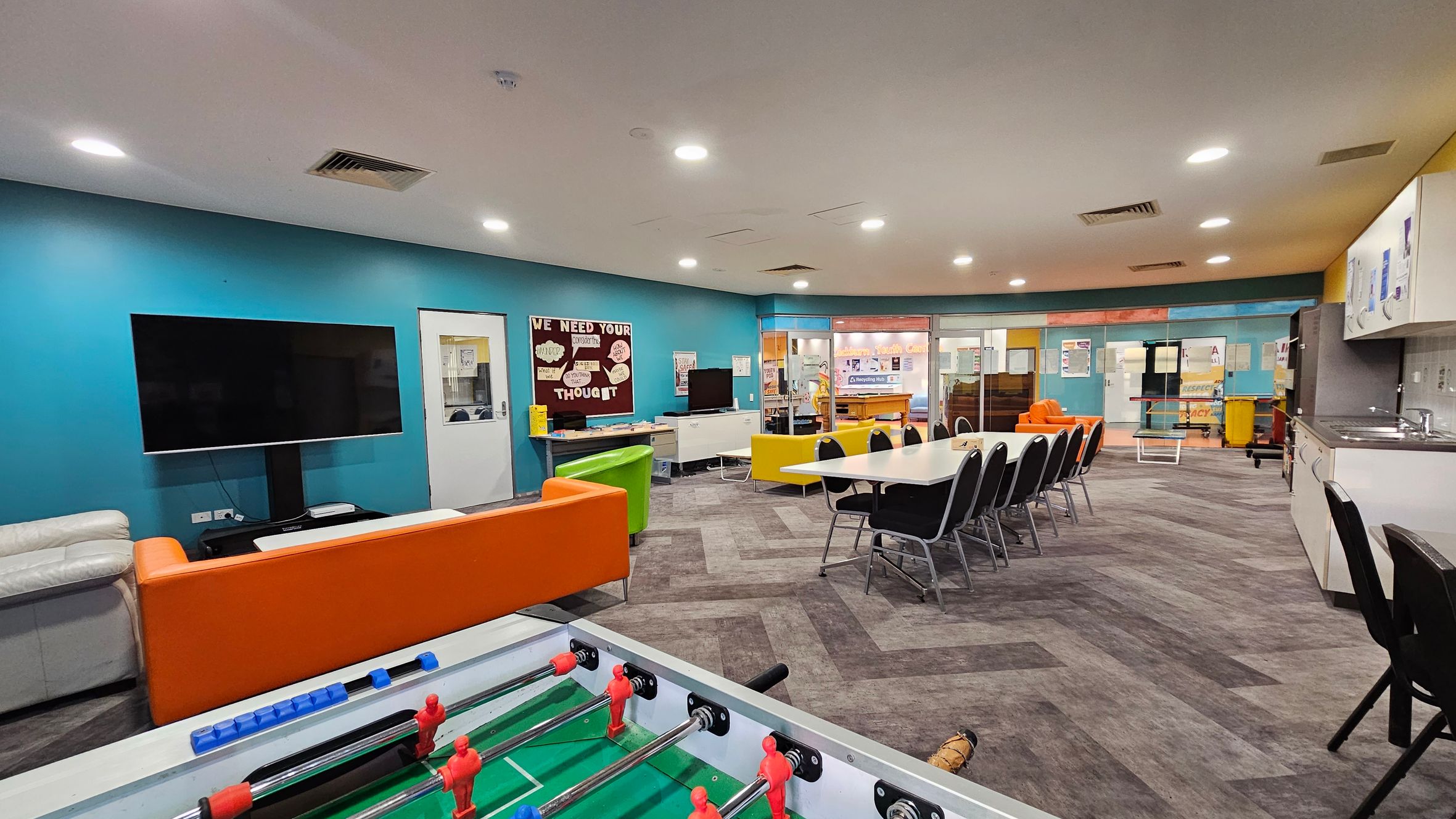 A spacious room with a foosball table in the foreground. On the left, there's a large TV mounted on a blue wall, with a white and orange sofa nearby. In the centre, a long table with black chairs is set up. The background features a bulletin board and a kitchenette area on the right, with a sink and cabinets. The floor has a patterned carpet, and the ceiling is equipped with recessed lighting. A spacious room with a foosball table in the foreground. On the left, there's a large TV mounted on a blue wall, with a white and orange sofa nearby. In the centre, a long table with black chairs is set up. The background features a bulletin board and a kitchenette area on the right, with a sink and cabinets. The floor has a patterned carpet, and the ceiling is equipped with recessed lighting.