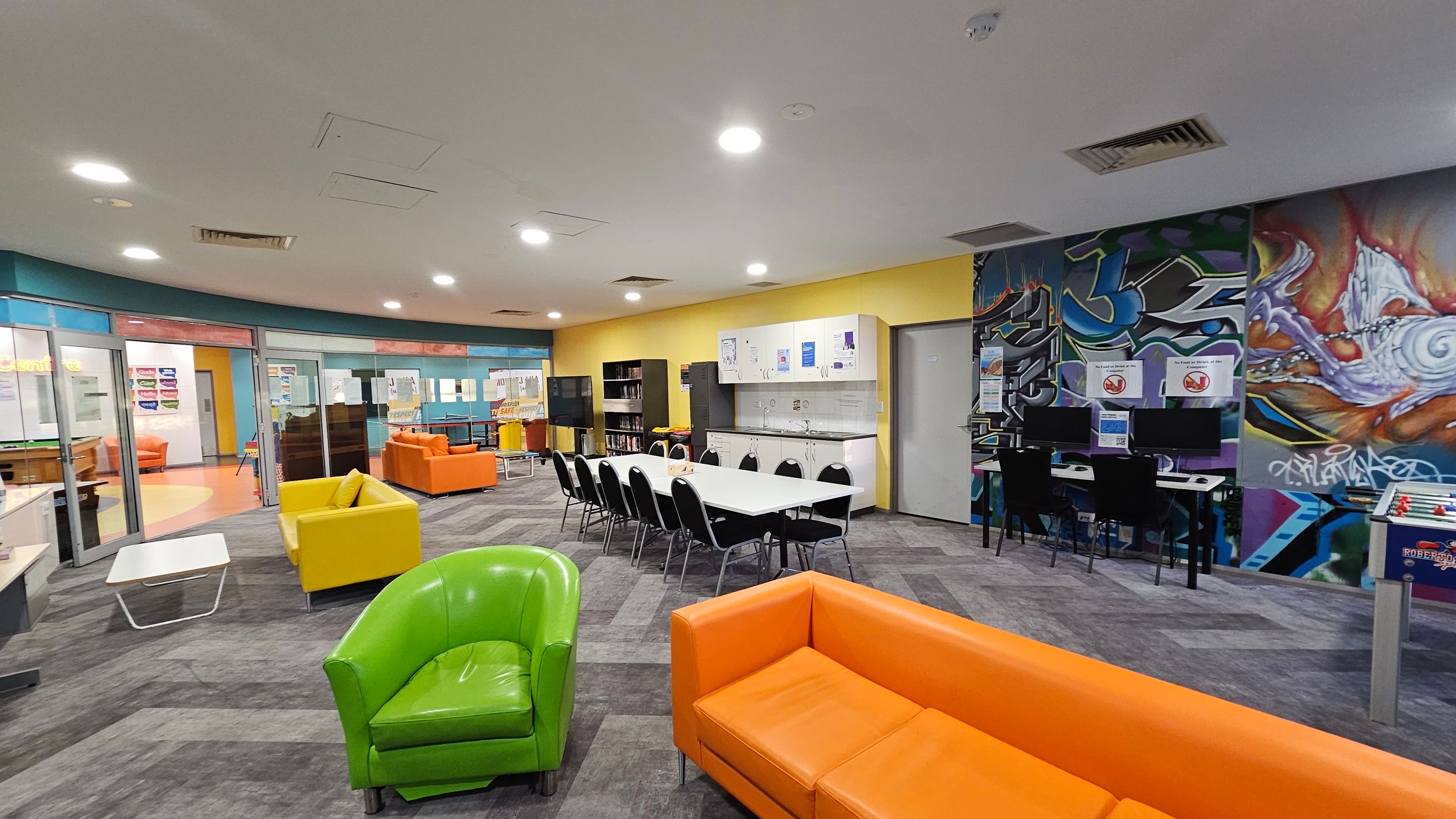 A spacious room with a modern design. On the left, there are glass doors leading to another area, with orange and yellow sofas visible. In the centre, a long white table with black chairs is set against a yellow wall with cabinets and a sink. On the right, a vibrant graffiti mural decorates the wall above computer stations. The floor is carpeted in a grey pattern, and the ceiling is white with recessed lighting. Brightly coloured sofas, including green and orange, are scattered throughout. A spacious room with a modern design. On the left, there are glass doors leading to another area, with orange and yellow sofas visible. In the centre, a long white table with black chairs is set against a yellow wall with cabinets and a sink. On the right, a vibrant graffiti mural decorates the wall above computer stations. The floor is carpeted in a grey pattern, and the ceiling is white with recessed lighting. Brightly coloured sofas, including green and orange, are scattered throughout.