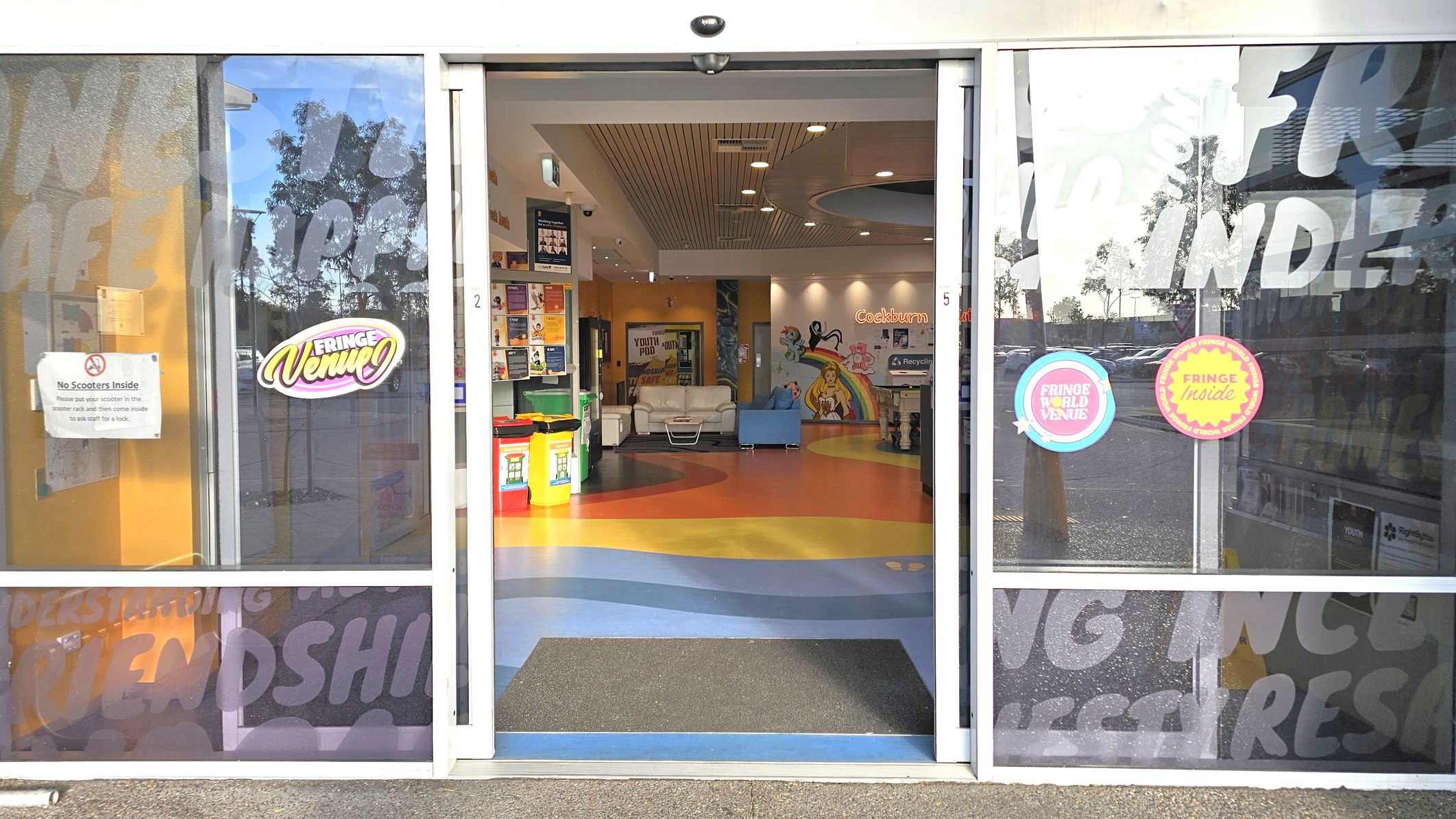 Sliding glass doors with signage, including "Fringe Venue" and "No Scooters Inside". Inside, a colourful floor with blue, yellow, and orange patterns leads to a seating area with white and blue couches. Walls are decorated with vibrant murals and posters. A recycling bin is visible on the right. The ceiling has recessed lighting and a wooden panel design. Reflections of trees and parked cars are seen on the glass. Sliding glass doors with signage, including "Fringe Venue" and "No Scooters Inside". Inside, a colourful floor with blue, yellow, and orange patterns leads to a seating area with white and blue couches. Walls are decorated with vibrant murals and posters. A recycling bin is visible on the right. The ceiling has recessed lighting and a wooden panel design. Reflections of trees and parked cars are seen on the glass.
