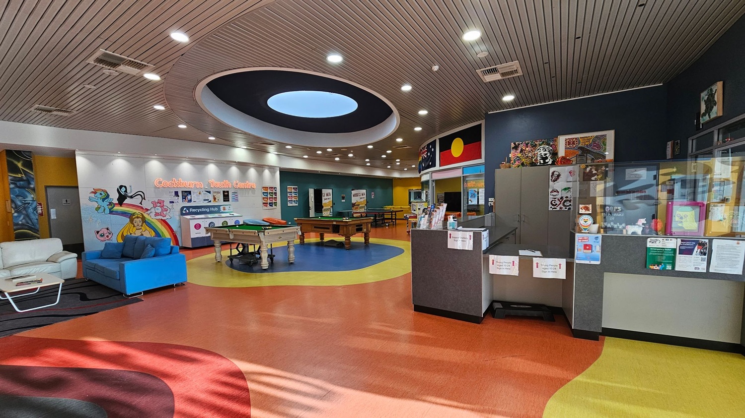 A colourful youth centre interior with a vibrant, patterned floor. On the left, there are white and blue sofas near a wall with cartoon murals and the words "Cockburn Youth Centre". In the centre, two pool tables are placed on a circular blue section of the floor. The right side features a reception area with a counter displaying brochures and posters. The ceiling is wooden with a large circular light fixture. Flags and artwork are displayed above the reception area. A colourful youth centre interior with a vibrant, patterned floor. On the left, there are white and blue sofas near a wall with cartoon murals and the words "Cockburn Youth Centre". In the centre, two pool tables are placed on a circular blue section of the floor. The right side features a reception area with a counter displaying brochures and posters. The ceiling is wooden with a large circular light fixture. Flags and artwork are displayed above the reception area.