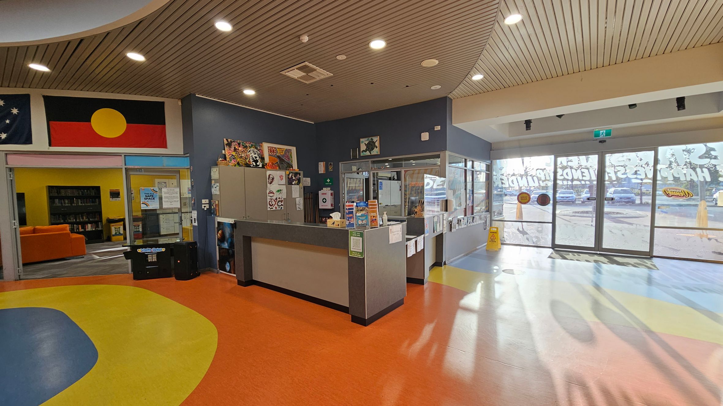 A reception area with a colourful floor featuring orange, yellow, and blue patterns. On the left, there is a library entrance with an orange sofa and bookshelves visible through glass doors. Above, flags are displayed on the wall. In the centre, a reception desk is adorned with various notices and items. To the right, large glass doors lead outside, with a wet floor sign nearby. The ceiling is wooden with recessed lighting. A reception area with a colourful floor featuring orange, yellow, and blue patterns. On the left, there is a library entrance with an orange sofa and bookshelves visible through glass doors. Above, flags are displayed on the wall. In the centre, a reception desk is adorned with various notices and items. To the right, large glass doors lead outside, with a wet floor sign nearby. The ceiling is wooden with recessed lighting.