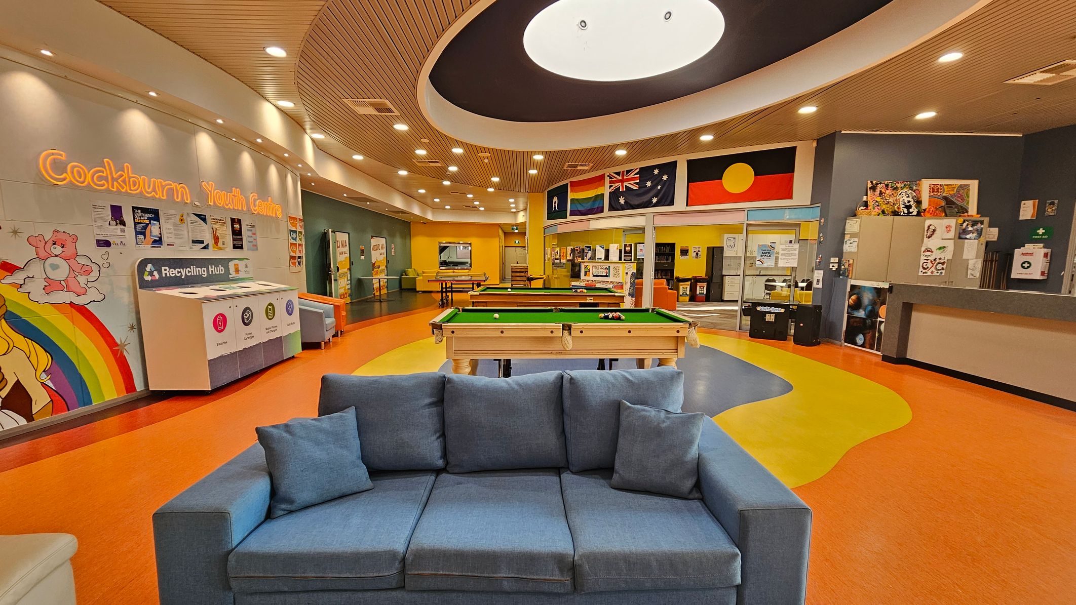 A vibrant indoor space with a blue sofa in the foreground. In the middle, there are pool tables on a colourful floor with orange, yellow, and blue patterns. The background features a wall with flags and a sign reading "Cockburn Youth Centre." On the left, there's a recycling hub and a mural with a rainbow and cartoon characters. The ceiling is curved with recessed lighting and a large circular light fixture. A vibrant indoor space with a blue sofa in the foreground. In the middle, there are pool tables on a colourful floor with orange, yellow, and blue patterns. The background features a wall with flags and a sign reading "Cockburn Youth Centre." On the left, there's a recycling hub and a mural with a rainbow and cartoon characters. The ceiling is curved with recessed lighting and a large circular light fixture.