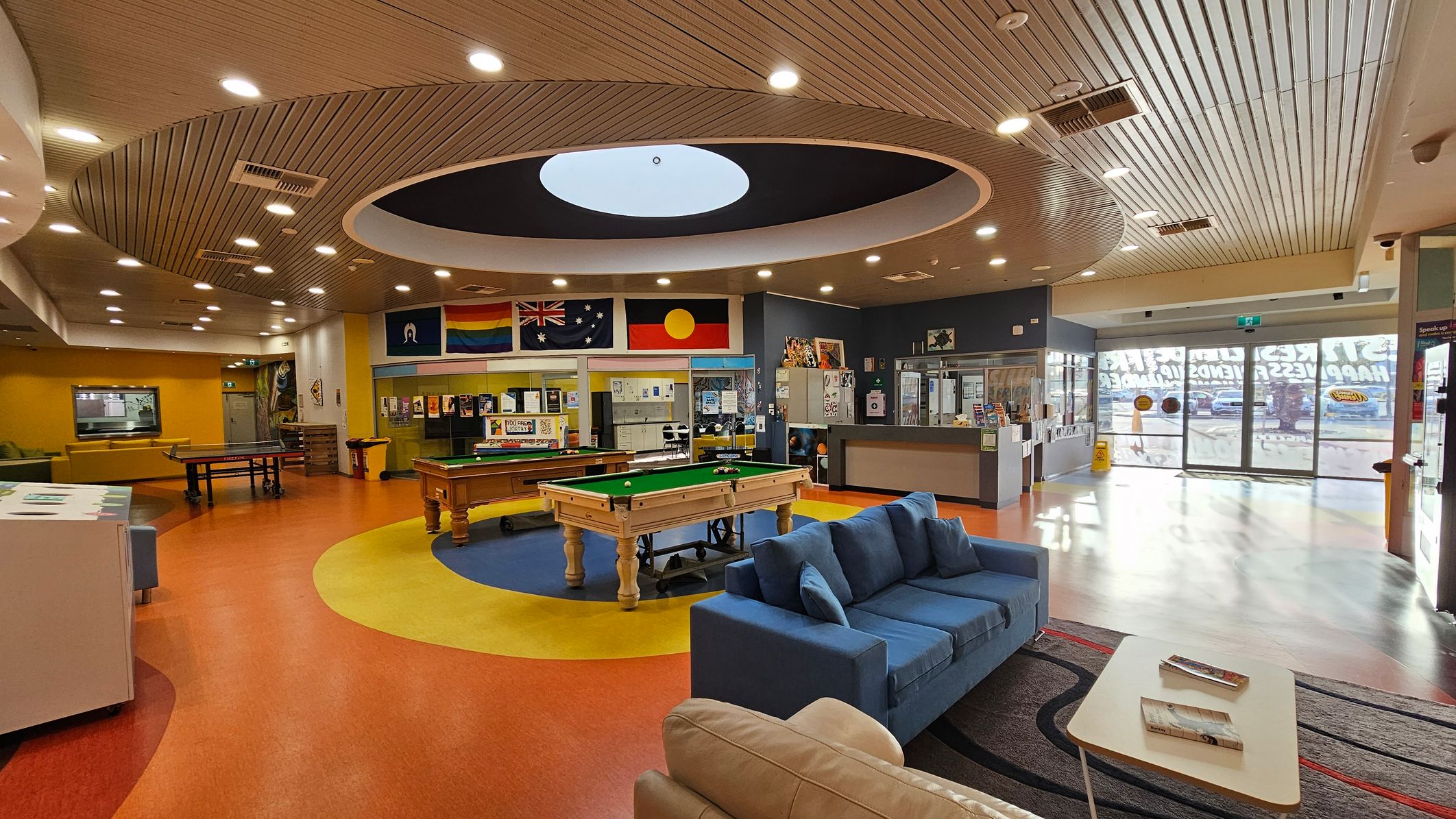 A spacious indoor recreational area with a circular ceiling design featuring recessed lighting. In the centre, there's a pool table on a blue and yellow circular carpet. To the left, a ping pong table and yellow seating area. On the right, a blue sofa and a small table with books. The back wall displays various flags and posters. The floor is a mix of red, yellow, and blue, and the entrance has large glass doors with signage. A spacious indoor recreational area with a circular ceiling design featuring recessed lighting. In the centre, there's a pool table on a blue and yellow circular carpet. To the left, a ping pong table and yellow seating area. On the right, a blue sofa and a small table with books. The back wall displays various flags and posters. The floor is a mix of red, yellow, and blue, and the entrance has large glass doors with signage.