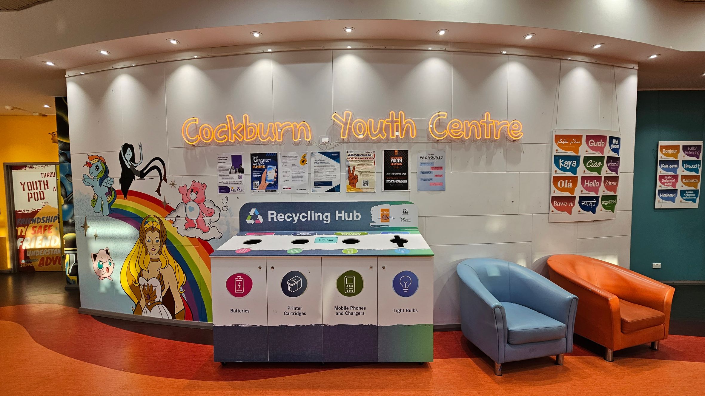 A colourful wall with the words "Cockburn Youth Centre" in neon lights. Below, a recycling hub with sections for batteries, printer cartridges, mobile phones, and light bulbs. To the left, a mural featuring cartoon characters and a rainbow. On the right, two chairs, one blue and one orange, sit on an orange floor. Posters with greetings in various languages are displayed on the right wall. The ceiling has recessed lighting. A colourful wall with the words "Cockburn Youth Centre" in neon lights. Below, a recycling hub with sections for batteries, printer cartridges, mobile phones, and light bulbs. To the left, a mural featuring cartoon characters and a rainbow. On the right, two chairs, one blue and one orange, sit on an orange floor. Posters with greetings in various languages are displayed on the right wall. The ceiling has recessed lighting.