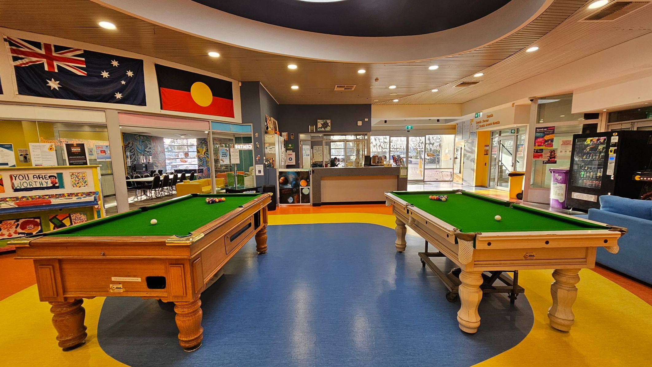Two pool tables with green felt tops are positioned in the centre of a room with a blue and yellow floor. Above, the ceiling is curved with recessed lighting. On the left wall, there are Australian and Aboriginal flags. Behind the pool tables, a glass partition reveals a room with chairs and tables. To the right, a vending machine and a blue sofa are visible. The background includes a reception area with a counter and glass doors leading outside. Two pool tables with green felt tops are positioned in the centre of a room with a blue and yellow floor. Above, the ceiling is curved with recessed lighting. On the left wall, there are Australian and Aboriginal flags. Behind the pool tables, a glass partition reveals a room with chairs and tables. To the right, a vending machine and a blue sofa are visible. The background includes a reception area with a counter and glass doors leading outside.