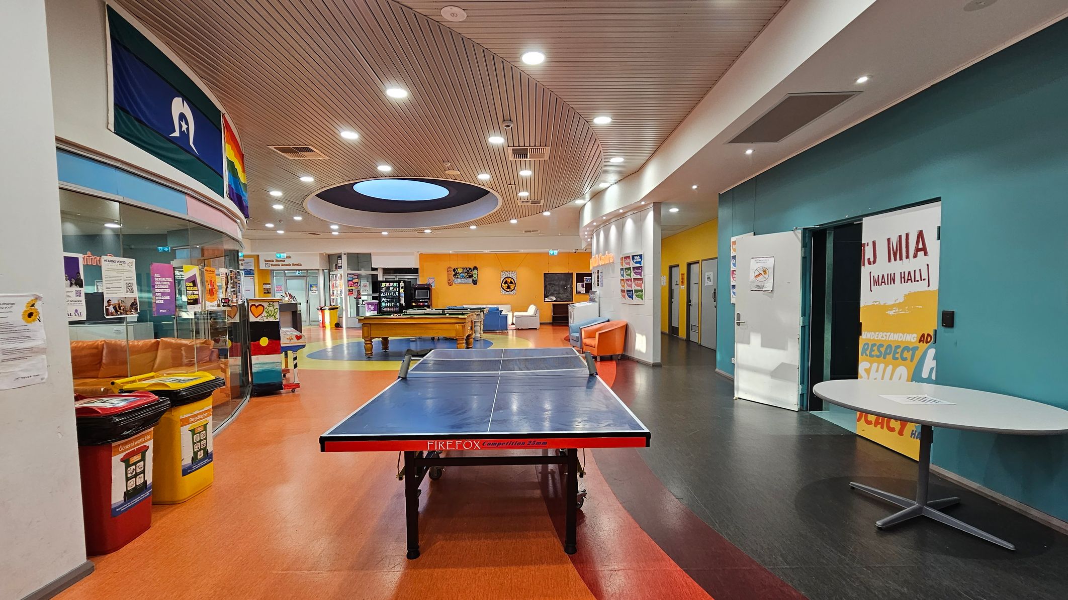 A spacious indoor recreational area with a ping pong table in the centre on an orange floor. To the left, there are colourful bins and a curved glass wall with posters and a rainbow flag. The right side features a teal wall with a door labelled "Main Hall" and a round table. In the background, there's a pool table, vending machines, and seating areas with couches. The ceiling is curved with recessed lighting and a circular skylight. A spacious indoor recreational area with a ping pong table in the centre on an orange floor. To the left, there are colourful bins and a curved glass wall with posters and a rainbow flag. The right side features a teal wall with a door labelled "Main Hall" and a round table. In the background, there's a pool table, vending machines, and seating areas with couches. The ceiling is curved with recessed lighting and a circular skylight.