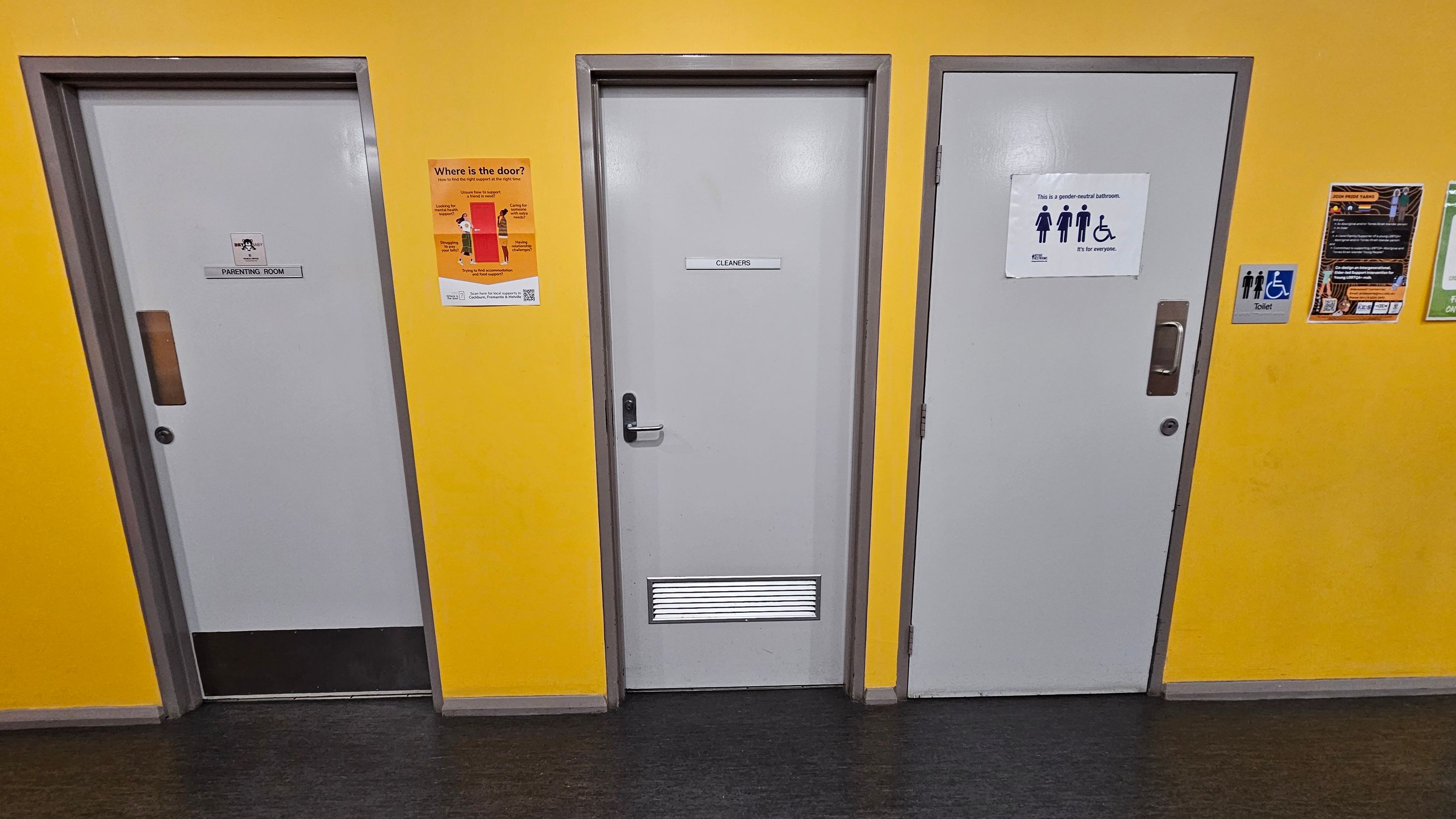 Three doors are set in a bright yellow wall. The left door is labelled "Parenting Room" with a small window. The middle door is marked "Cleaners" and has a vent at the bottom. The right door is a gender-neutral bathroom, indicated by a sign with symbols for male, female, and wheelchair access. Various informational posters are on the wall, including one about door locations. The floor is dark grey, and the ceiling is not visible. Three doors are set in a bright yellow wall. The left door is labelled "Parenting Room" with a small window. The middle door is marked "Cleaners" and has a vent at the bottom. The right door is a gender-neutral bathroom, indicated by a sign with symbols for male, female, and wheelchair access. Various informational posters are on the wall, including one about door locations. The floor is dark grey, and the ceiling is not visible.
