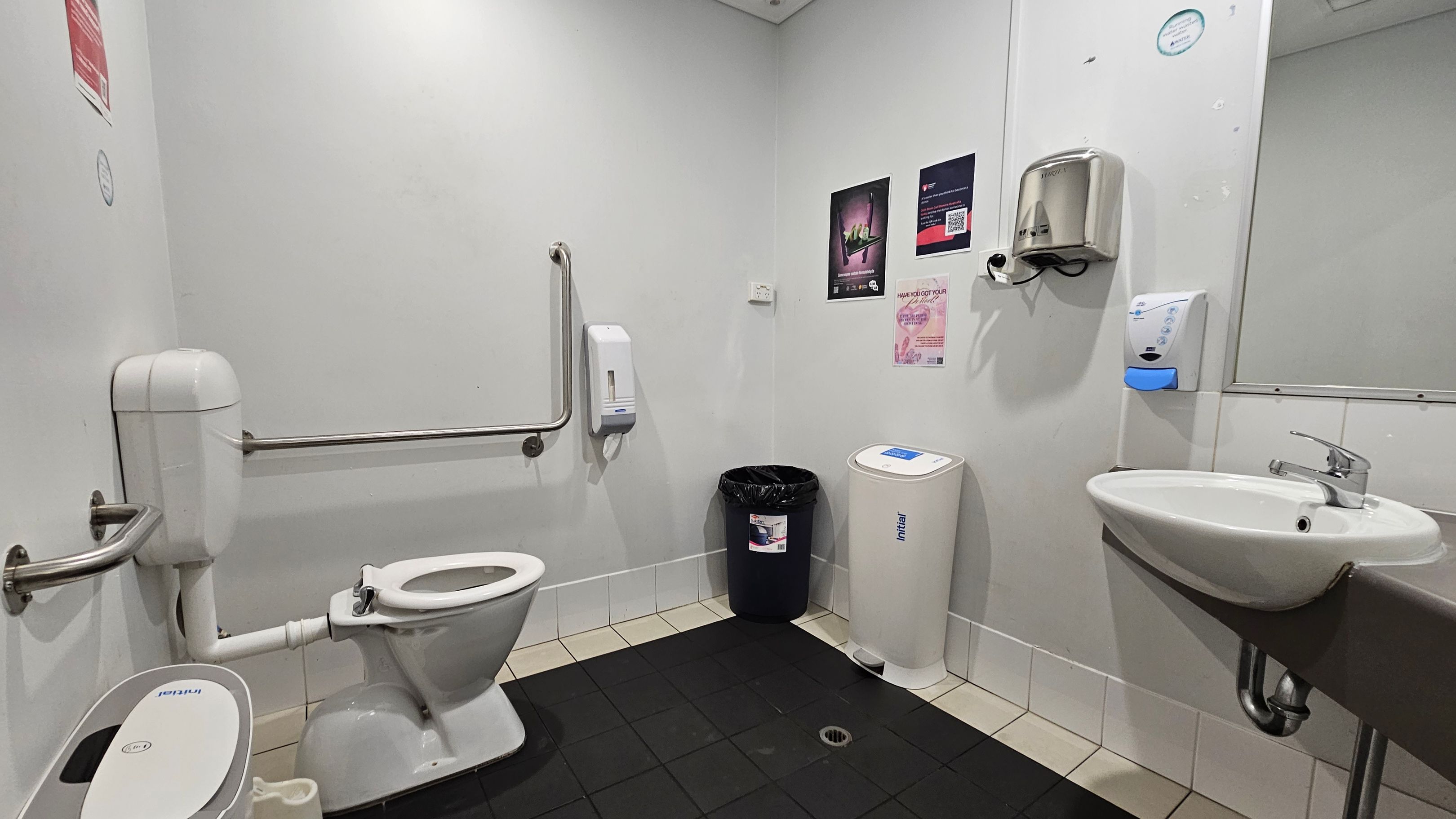 A public restroom with a white toilet on the left, equipped with a support rail. A black bin and a white sanitary bin are against the back wall. The right side features a wall-mounted sink with a silver tap, a soap dispenser, and a hand dryer above. The walls are adorned with posters. The floor is tiled in black and beige, and the ceiling is plain white with recessed lighting. A public restroom with a white toilet on the left, equipped with a support rail. A black bin and a white sanitary bin are against the back wall. The right side features a wall-mounted sink with a silver tap, a soap dispenser, and a hand dryer above. The walls are adorned with posters. The floor is tiled in black and beige, and the ceiling is plain white with recessed lighting.