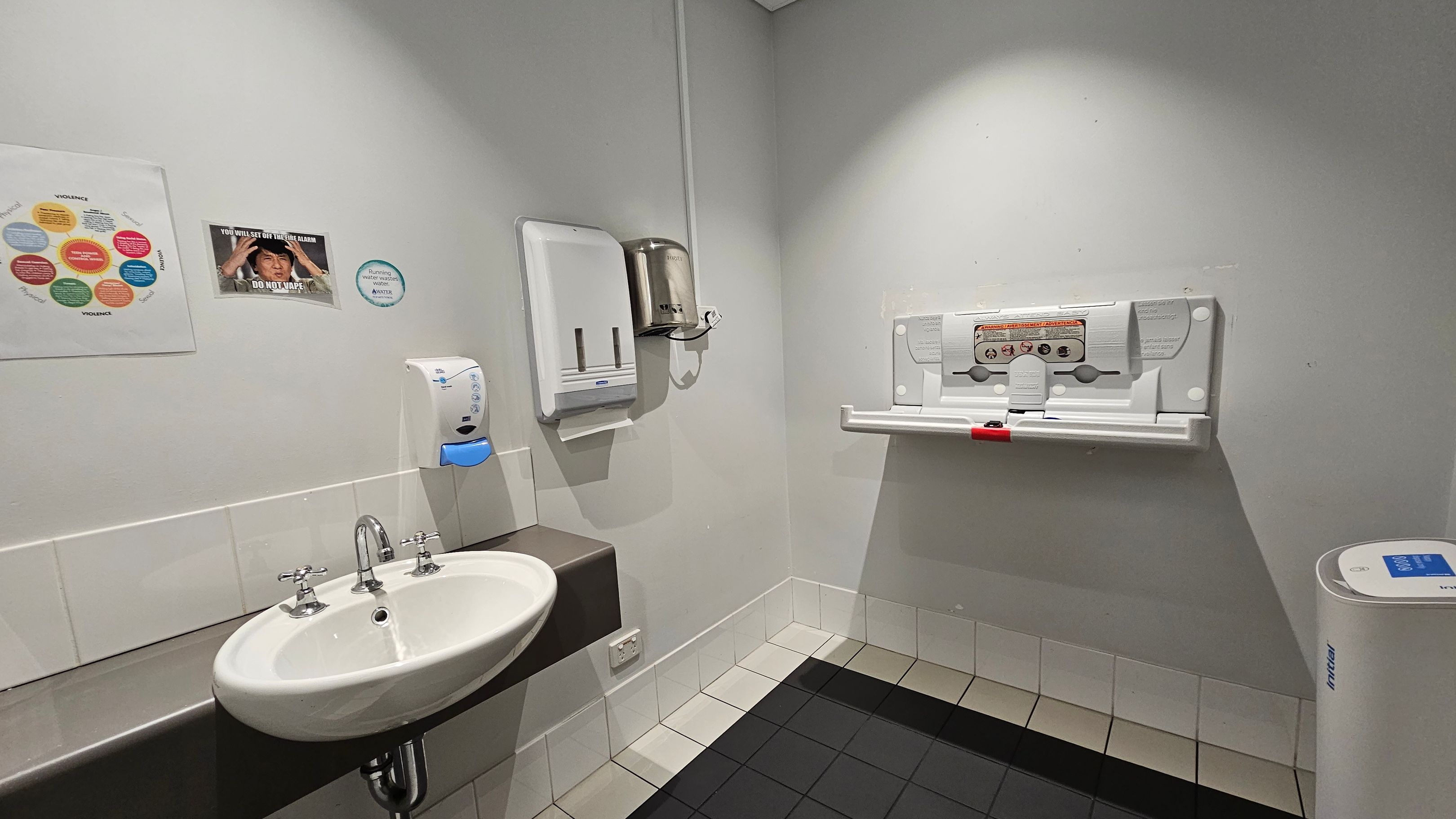 A public restroom with a white sink and silver taps on the left, mounted on a brown counter. Above the sink are a soap dispenser and a paper towel holder. On the wall, there are posters and a circular sign. To the right, a baby changing station is attached to the wall. The floor is tiled in black and light colours, and the walls are light-coloured. A white bin is in the corner on the right. The ceiling is plain and light-coloured. A public restroom with a white sink and silver taps on the left, mounted on a brown counter. Above the sink are a soap dispenser and a paper towel holder. On the wall, there are posters and a circular sign. To the right, a baby changing station is attached to the wall. The floor is tiled in black and light colours, and the walls are light-coloured. A white bin is in the corner on the right. The ceiling is plain and light-coloured.