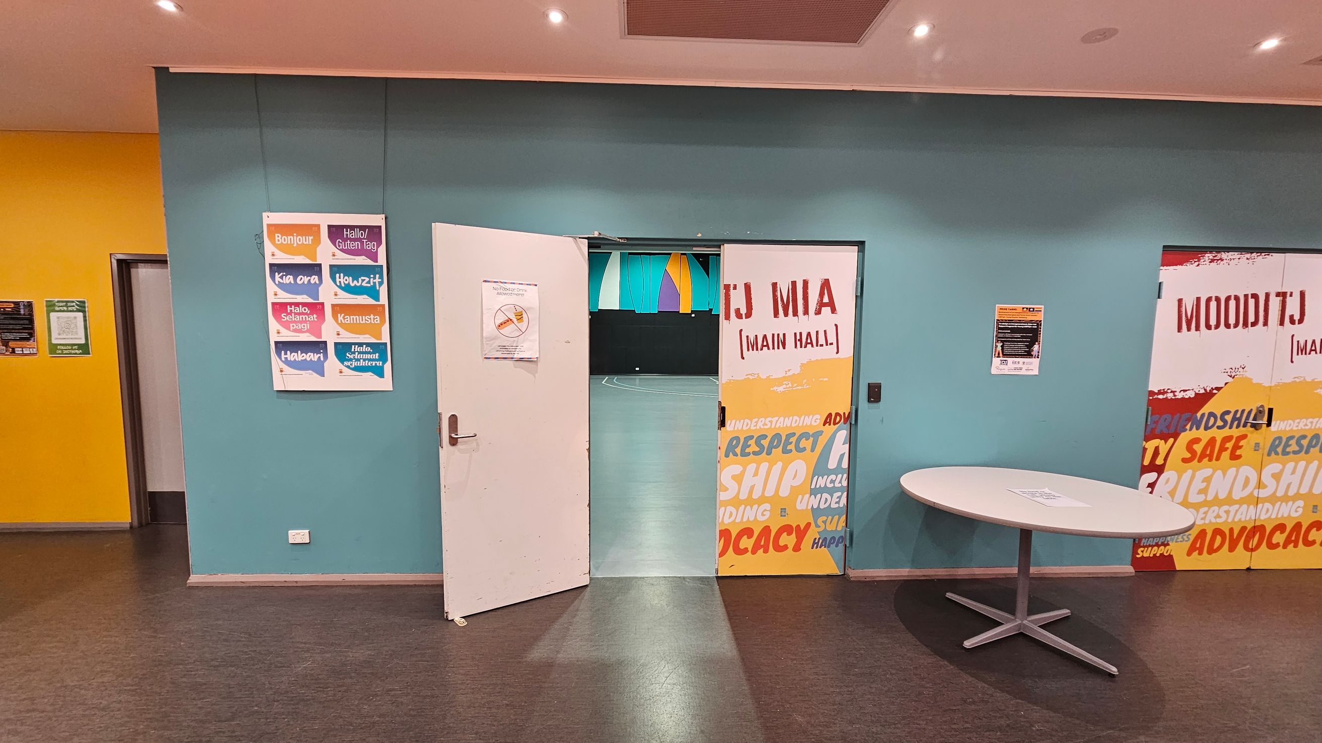 A room with a blue wall featuring a partially open door leading to a hall. The door has a sign with "TJ MIA (MAIN HALL)" and colourful words like "RESPECT" and "FRIENDSHIP." To the left, a poster displays greetings in various languages. On the right, a round table with a paper on it. The floor is dark, and the ceiling has recessed lighting. The left wall is yellow with a small doorway. A room with a blue wall featuring a partially open door leading to a hall. The door has a sign with "TJ MIA (MAIN HALL)" and colourful words like "RESPECT" and "FRIENDSHIP." To the left, a poster displays greetings in various languages. On the right, a round table with a paper on it. The floor is dark, and the ceiling has recessed lighting. The left wall is yellow with a small doorway.
