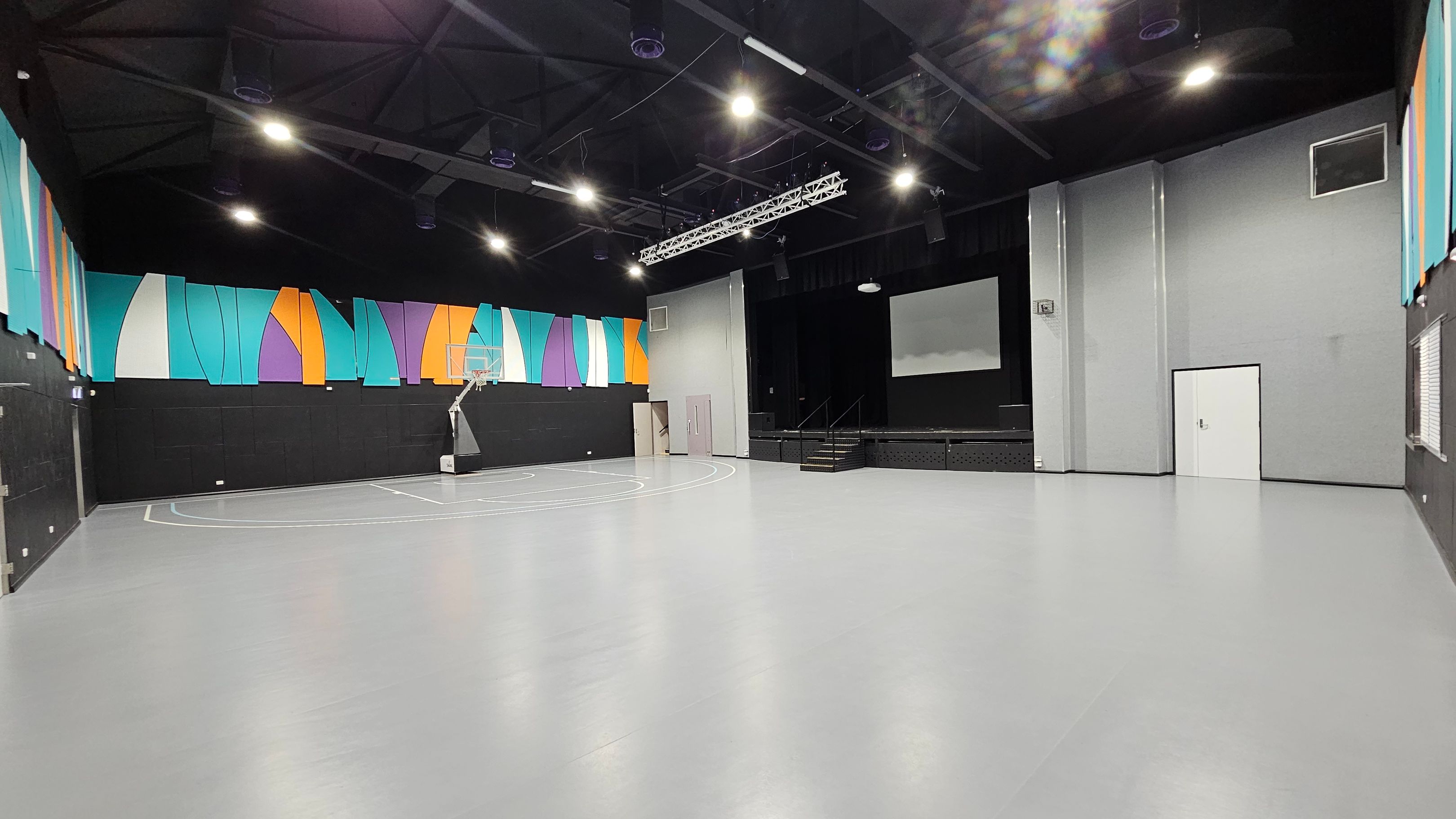 A spacious indoor gymnasium with a smooth grey floor and a high, dark ceiling. On the left, colourful geometric panels in blue, orange, and purple adorn the upper walls. A basketball hoop is positioned near the left wall. In the centre, a stage with stairs is visible, featuring a large screen above it. On the right, a plain grey wall with a white door and a small window. Bright overhead lights illuminate the space. A spacious indoor gymnasium with a smooth grey floor and a high, dark ceiling. On the left, colourful geometric panels in blue, orange, and purple adorn the upper walls. A basketball hoop is positioned near the left wall. In the centre, a stage with stairs is visible, featuring a large screen above it. On the right, a plain grey wall with a white door and a small window. Bright overhead lights illuminate the space.