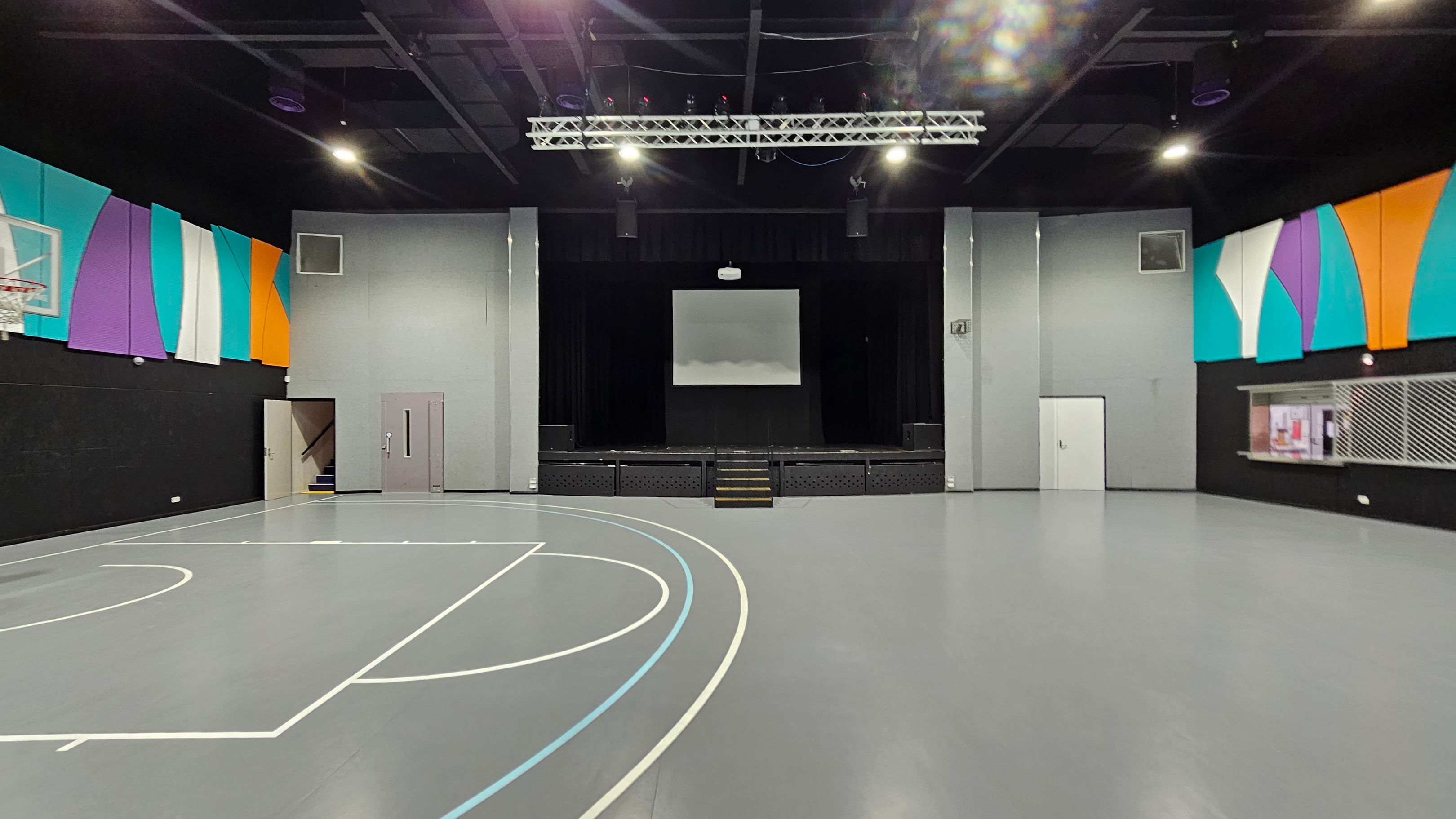 A spacious indoor gymnasium with a grey floor featuring basketball court lines. On the left, a basketball hoop is mounted on a wall adorned with colourful panels in teal, purple, white, and orange. In the centre, a stage with a screen above it is flanked by grey walls. On the right, a serving counter with a metal grille is set into the wall, which also has colourful panels. The ceiling is black with exposed beams and lighting fixtures. A spacious indoor gymnasium with a grey floor featuring basketball court lines. On the left, a basketball hoop is mounted on a wall adorned with colourful panels in teal, purple, white, and orange. In the centre, a stage with a screen above it is flanked by grey walls. On the right, a serving counter with a metal grille is set into the wall, which also has colourful panels. The ceiling is black with exposed beams and lighting fixtures.