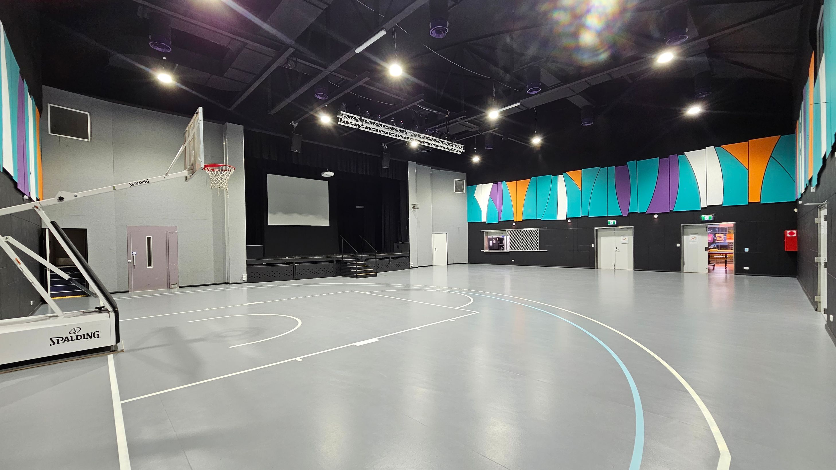 A spacious indoor gymnasium with a grey floor marked for basketball. On the left, a basketball hoop with a "Spalding" logo is visible. The back wall features a stage with a large screen and steps leading up to it. The right wall is adorned with colourful geometric panels in teal, orange, and purple. Several doors are present, including an open one showing a glimpse of another room. The ceiling is high with exposed beams and multiple lights. A spacious indoor gymnasium with a grey floor marked for basketball. On the left, a basketball hoop with a "Spalding" logo is visible. The back wall features a stage with a large screen and steps leading up to it. The right wall is adorned with colourful geometric panels in teal, orange, and purple. Several doors are present, including an open one showing a glimpse of another room. The ceiling is high with exposed beams and multiple lights.