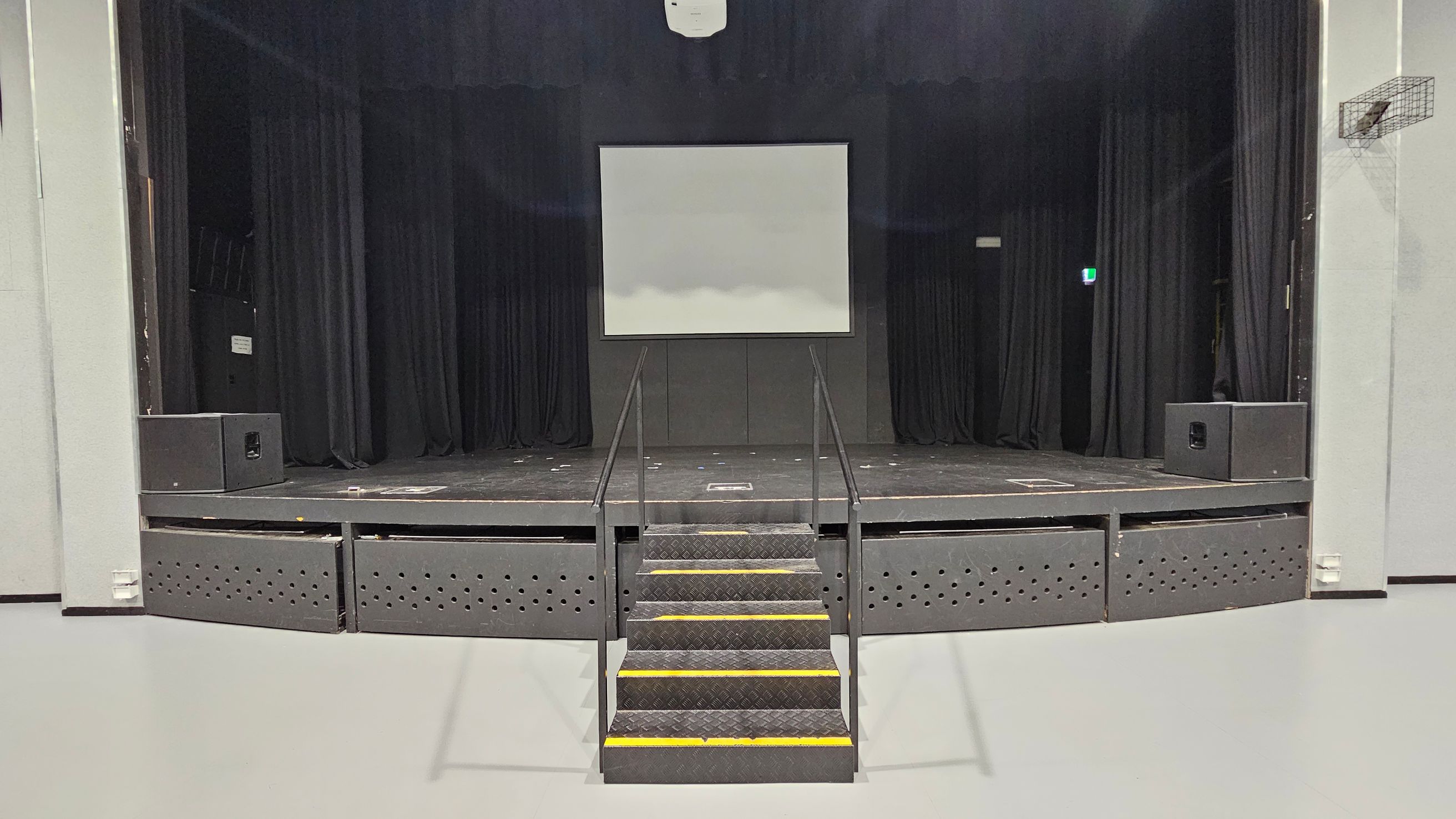 A stage with a central staircase leading up to it, featuring black railings. The stage has a large white screen in the middle, flanked by black curtains. Two large speakers are positioned on either side of the stage. The floor is light grey, and the ceiling is not visible. A small exit sign is illuminated on the right side. A stage with a central staircase leading up to it, featuring black railings. The stage has a large white screen in the middle, flanked by black curtains. Two large speakers are positioned on either side of the stage. The floor is light grey, and the ceiling is not visible. A small exit sign is illuminated on the right side.