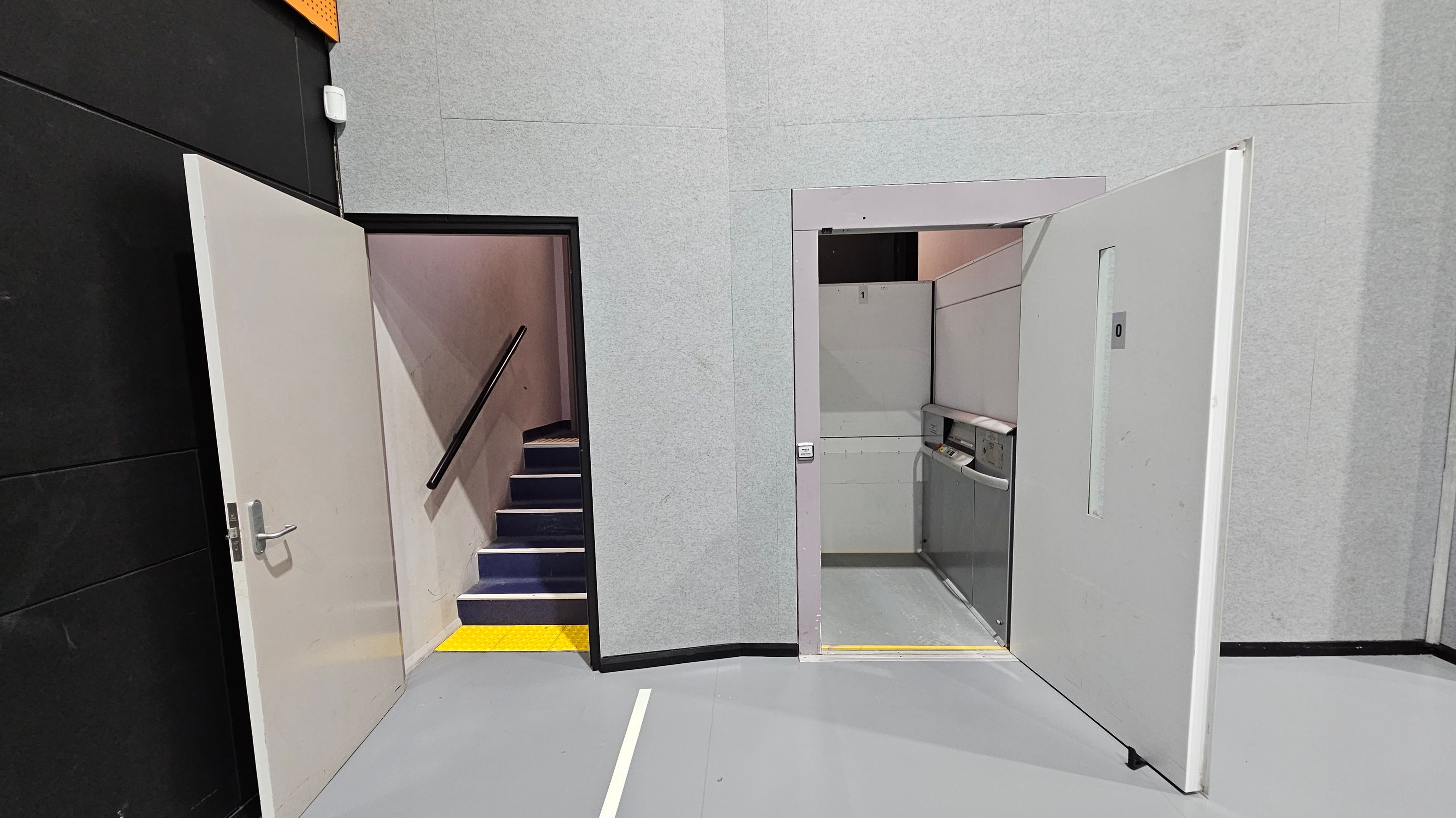 On the left, a door is open, revealing a staircase with a black handrail and blue steps, bordered by a yellow strip. On the right, another open door leads to a small lift with control panels visible. The walls are grey, and the floor is a smooth, light grey surface. The ceiling is not visible. On the left, a door is open, revealing a staircase with a black handrail and blue steps, bordered by a yellow strip. On the right, another open door leads to a small lift with control panels visible. The walls are grey, and the floor is a smooth, light grey surface. The ceiling is not visible.
