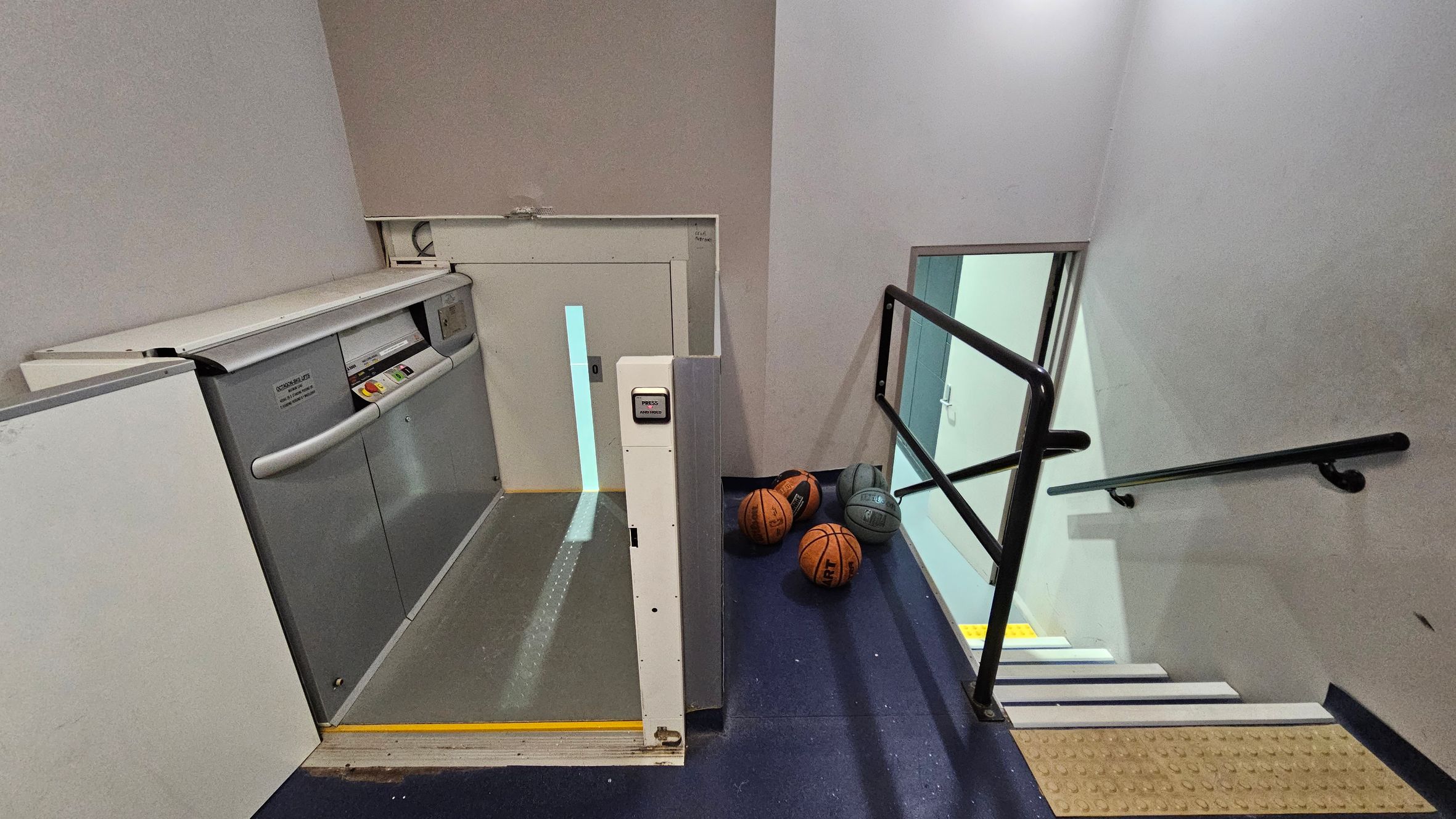 A stairwell with a platform lift on the left, featuring a control panel and safety rails. On the right, a set of stairs descends with black handrails. At the bottom of the stairs, several basketballs are scattered on the floor. The walls are light-coloured, and the floor is dark. The ceiling is not visible. A stairwell with a platform lift on the left, featuring a control panel and safety rails. On the right, a set of stairs descends with black handrails. At the bottom of the stairs, several basketballs are scattered on the floor. The walls are light-coloured, and the floor is dark. The ceiling is not visible.